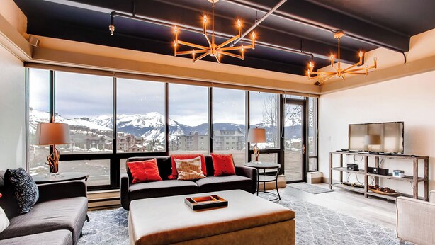 A spacious living room with a large window offering a mountain view, a modern chandelier, and a cozy seating area with a sofa and ottoman.