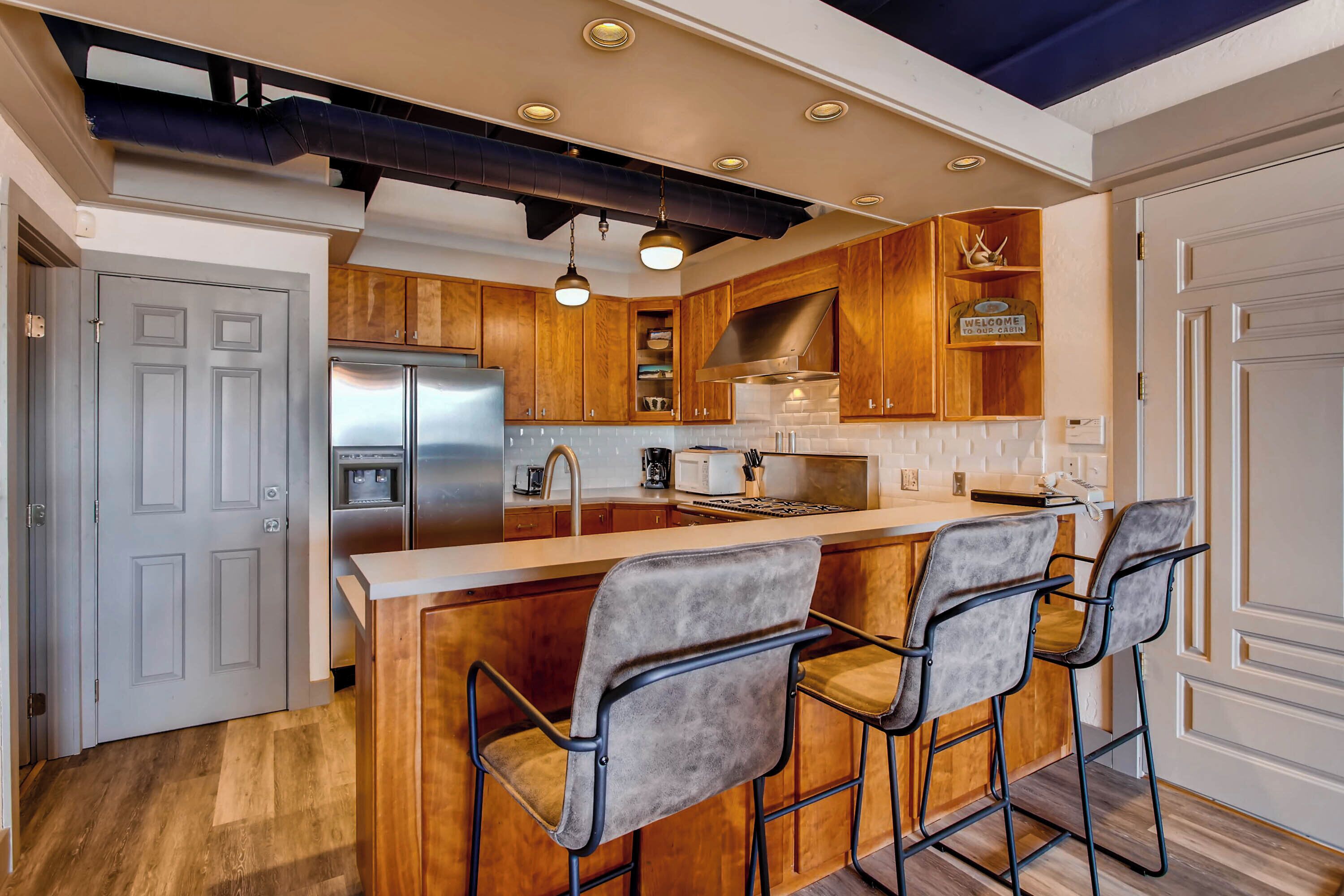 A modern kitchen with wooden cabinets, a stainless steel refrigerator, a built-in oven, and a bar area with bar stools.