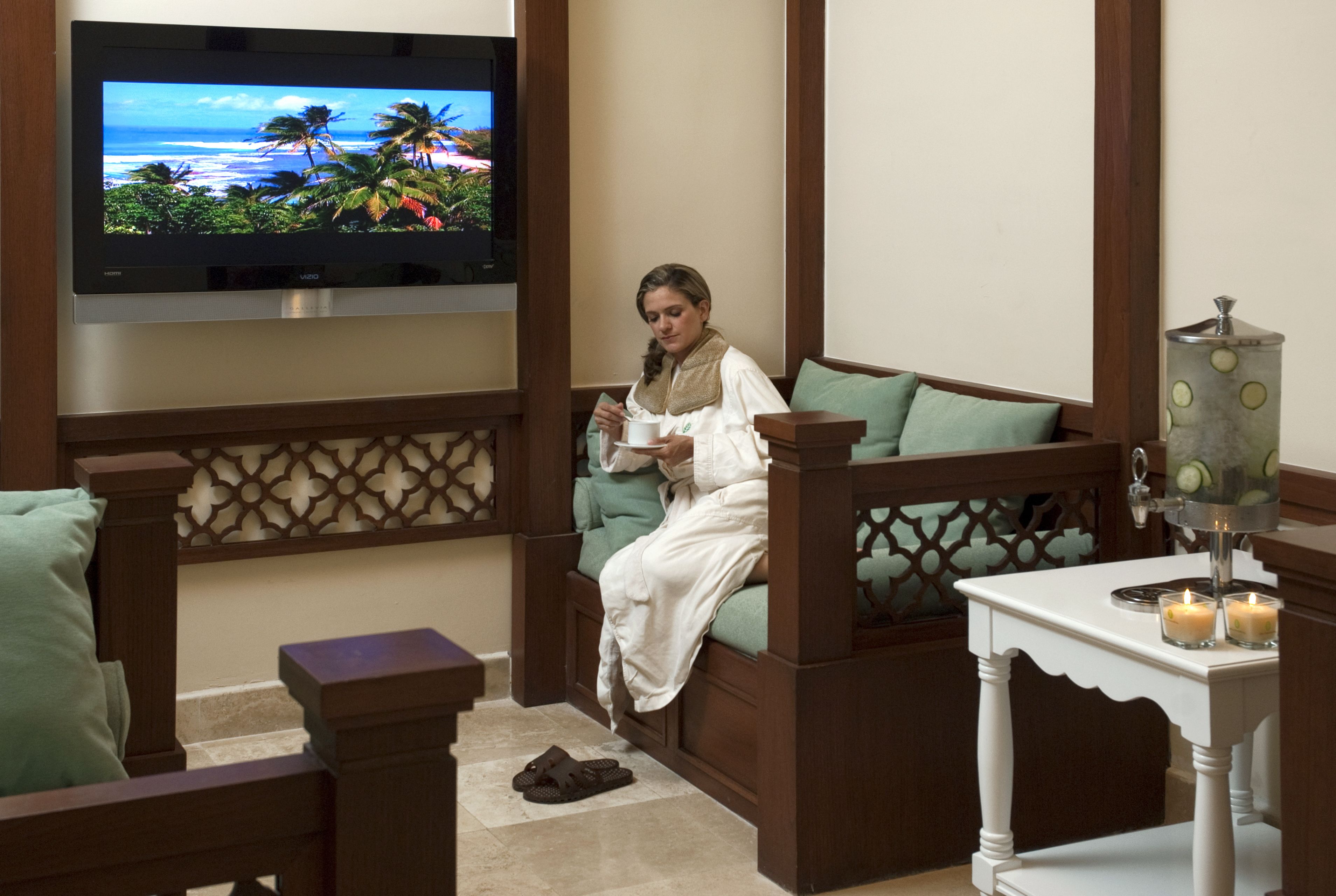 A person sitting on a sofa in a room with a TV displaying a tropical beach scene, a small table with candles, and a decorative container.