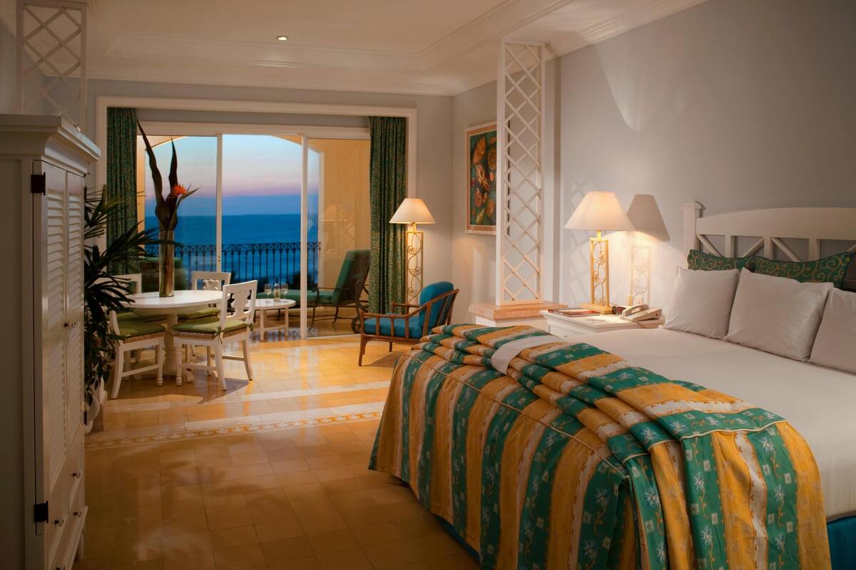 A hotel room with a bed, a desk with a chair, a balcony with a view, and a nightstand with lamps.