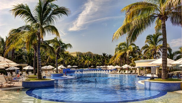 Uma piscina grande com espreguiçadeiras e guarda-sóis, cercada por palmeiras e um edifício ao fundo.