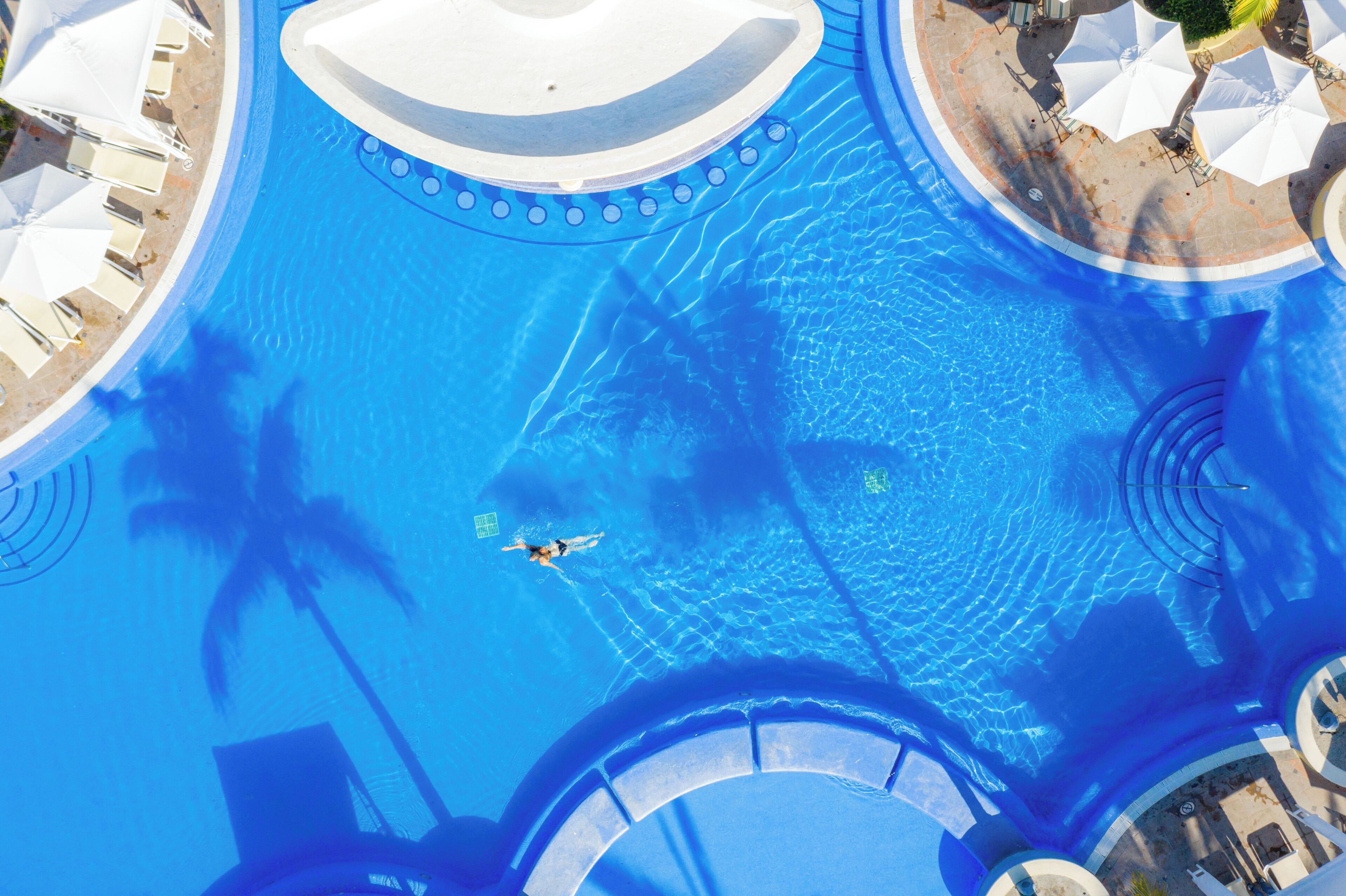 An aerial view of a swimming pool with a person swimming, surrounded by lounge chairs and umbrellas.