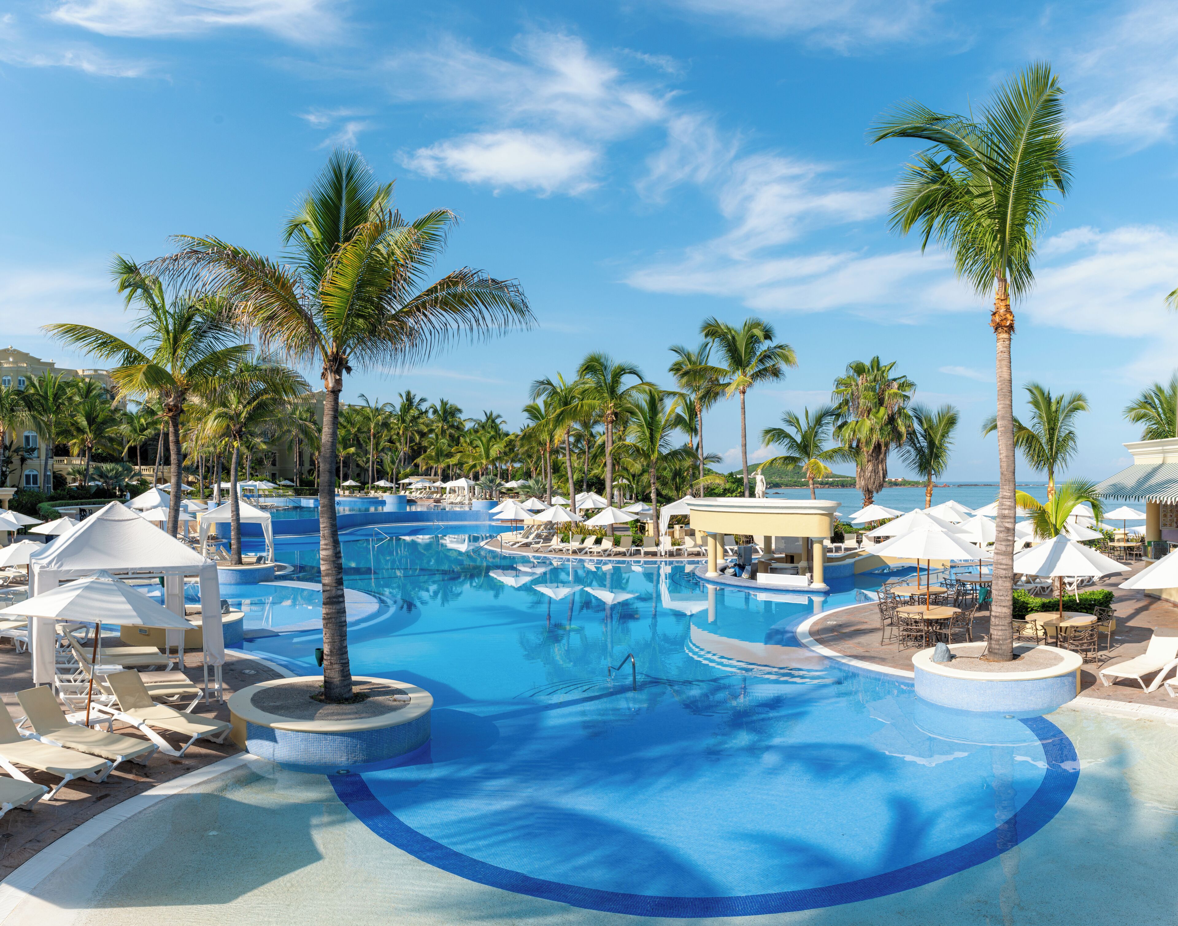 Un área de piscina en un complejo turístico con palmeras, sillones de playa y sombrillas.