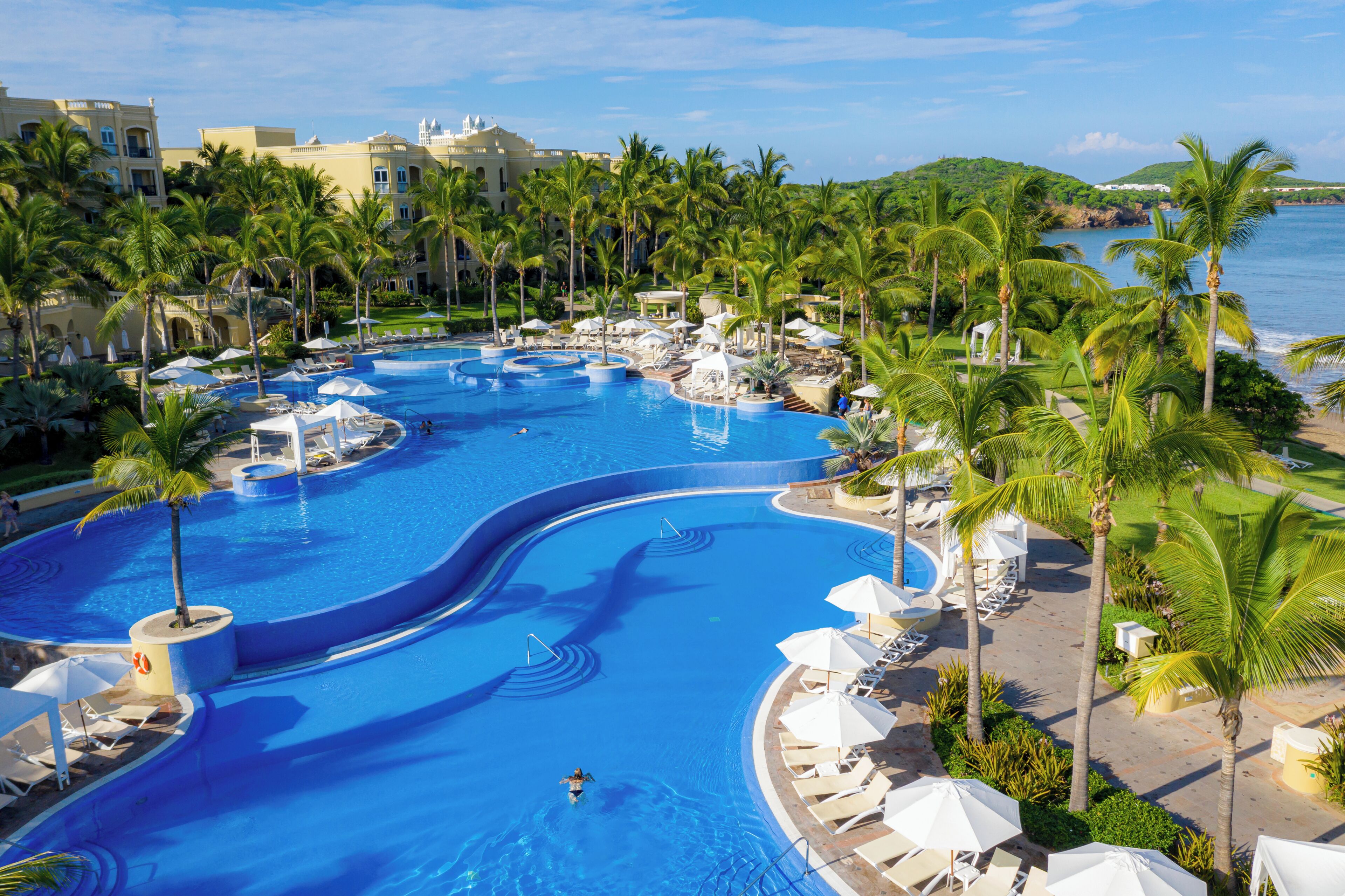 Piscina de resort con varias secciones, rodeada de palmeras y sillas de descanso.