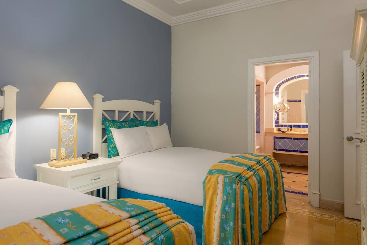 A hotel room with two beds, a nightstand, a lamp, and a bathroom visible through an open door.