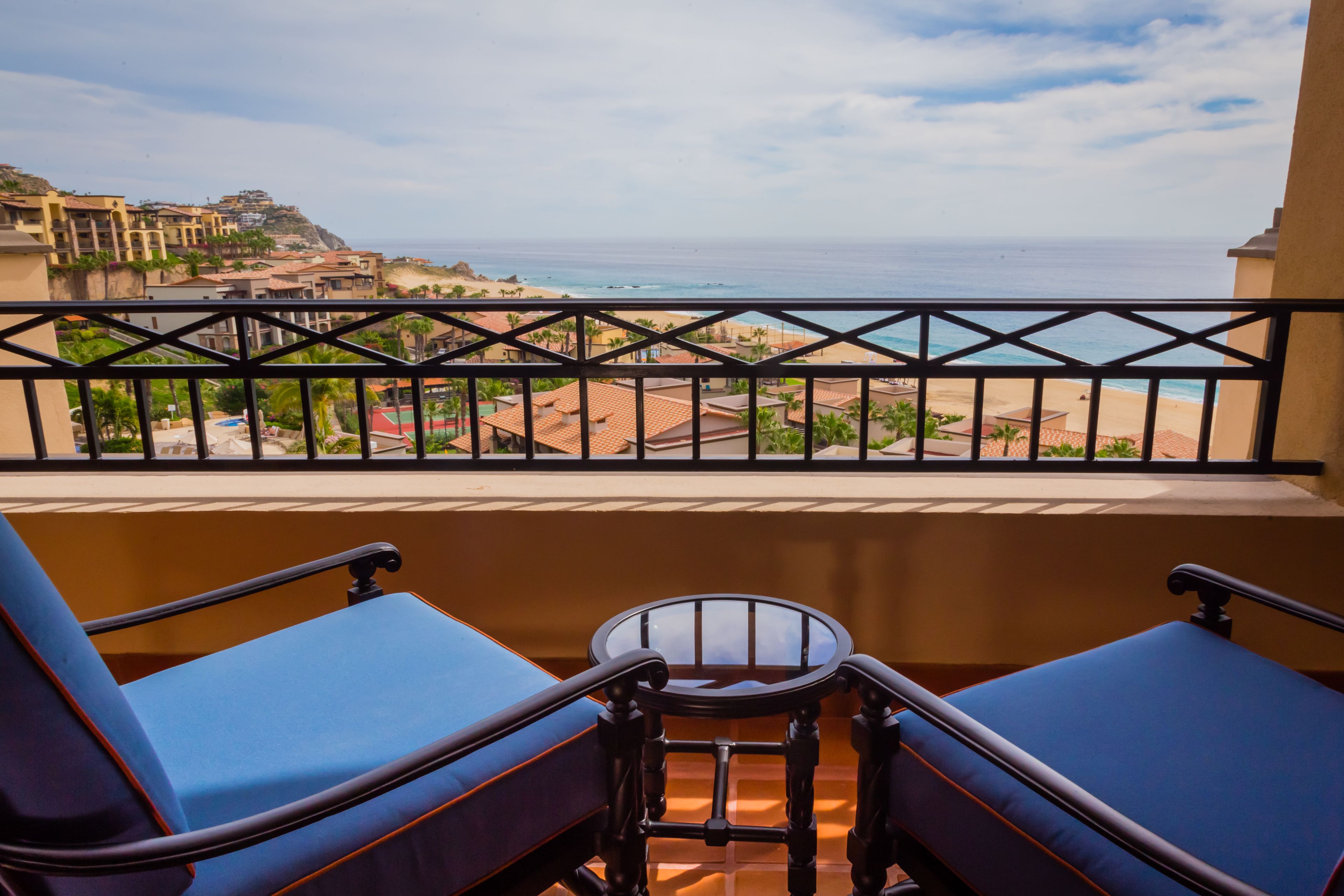 A balcony with two blue cushioned chairs and a small table, overlooking a beach and a coastal town.