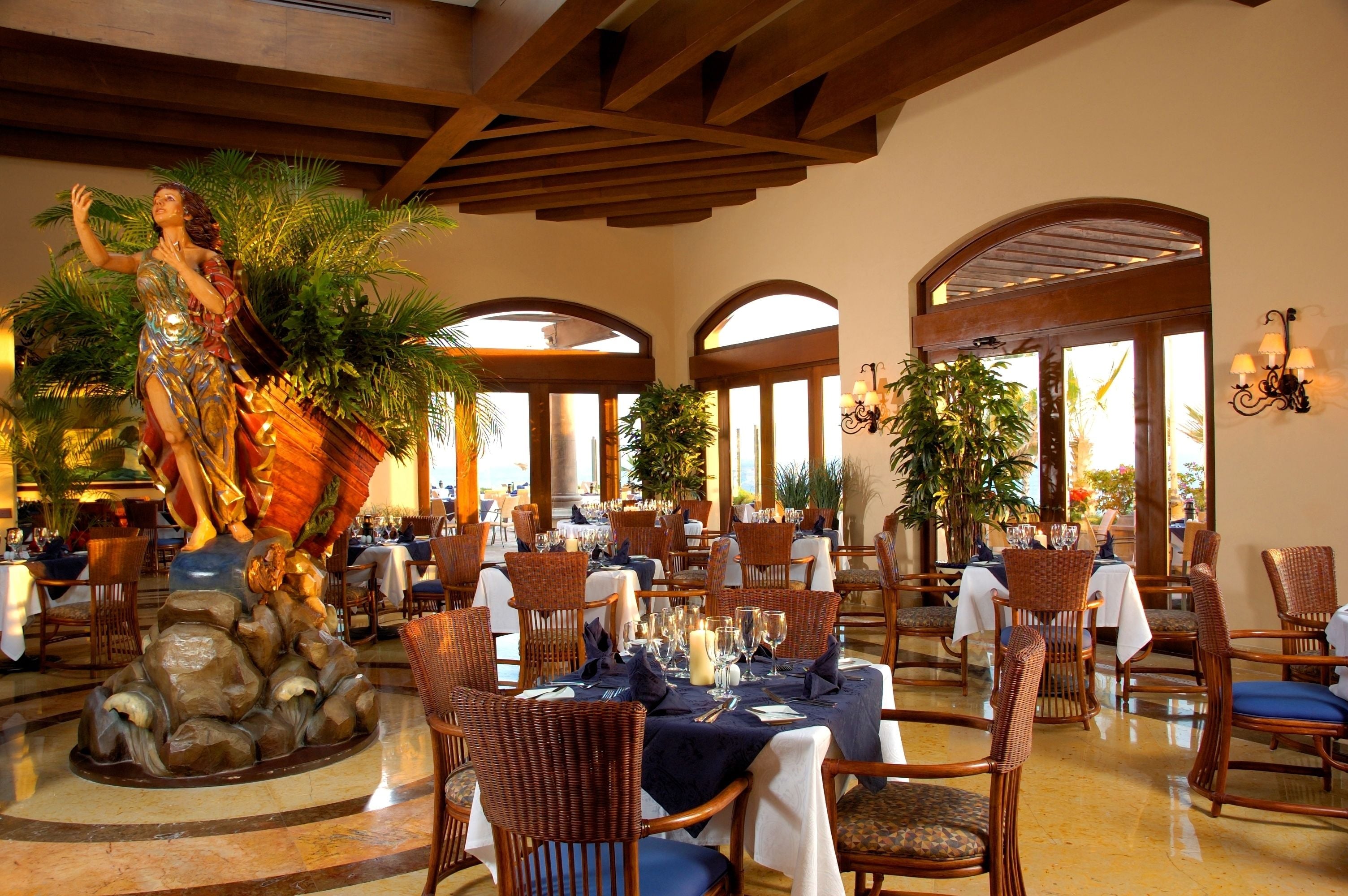 A restaurant with tables set for dining, a statue, and arched windows.