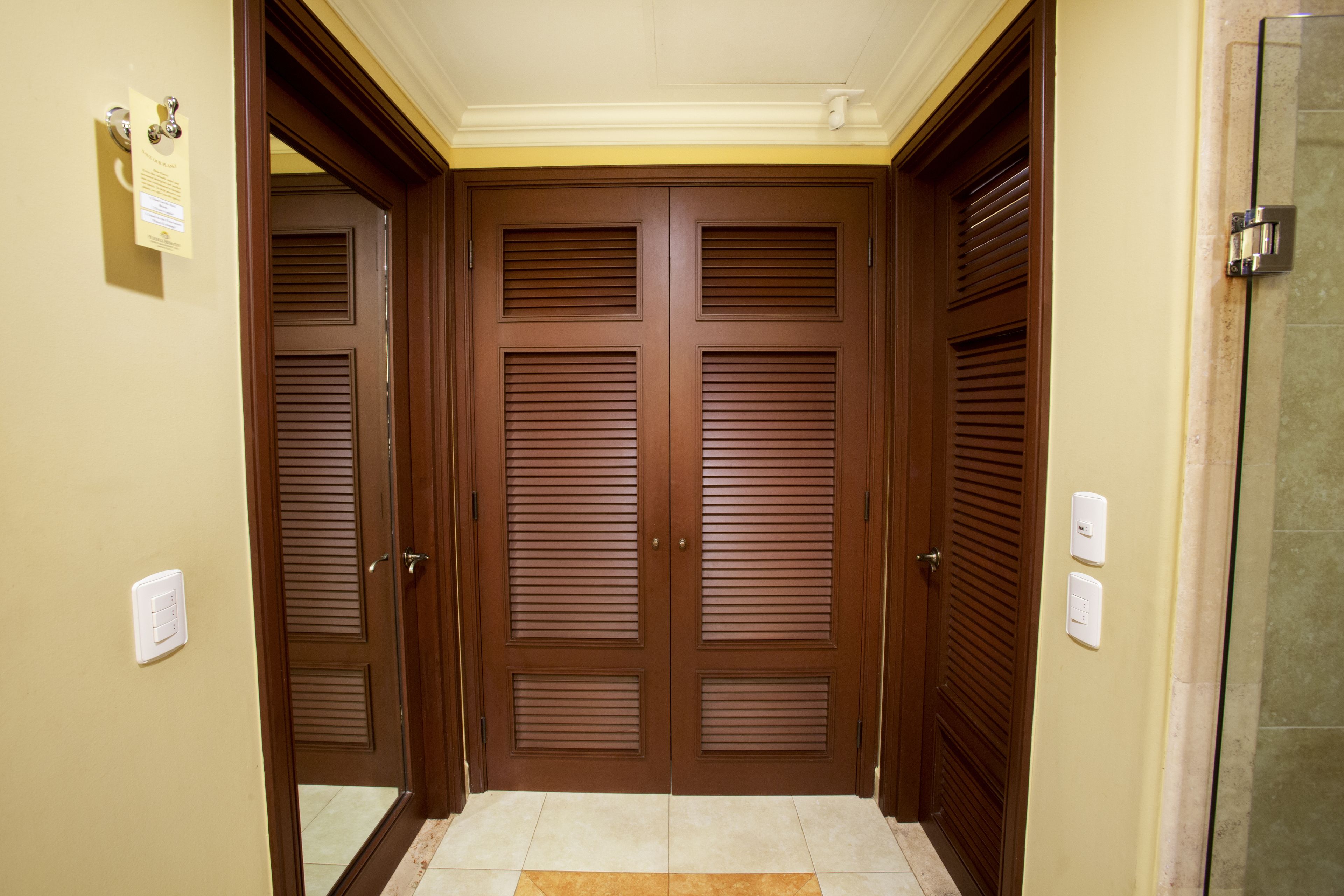 A hallway with double doors, a tiled floor, and a light switch on the wall.
