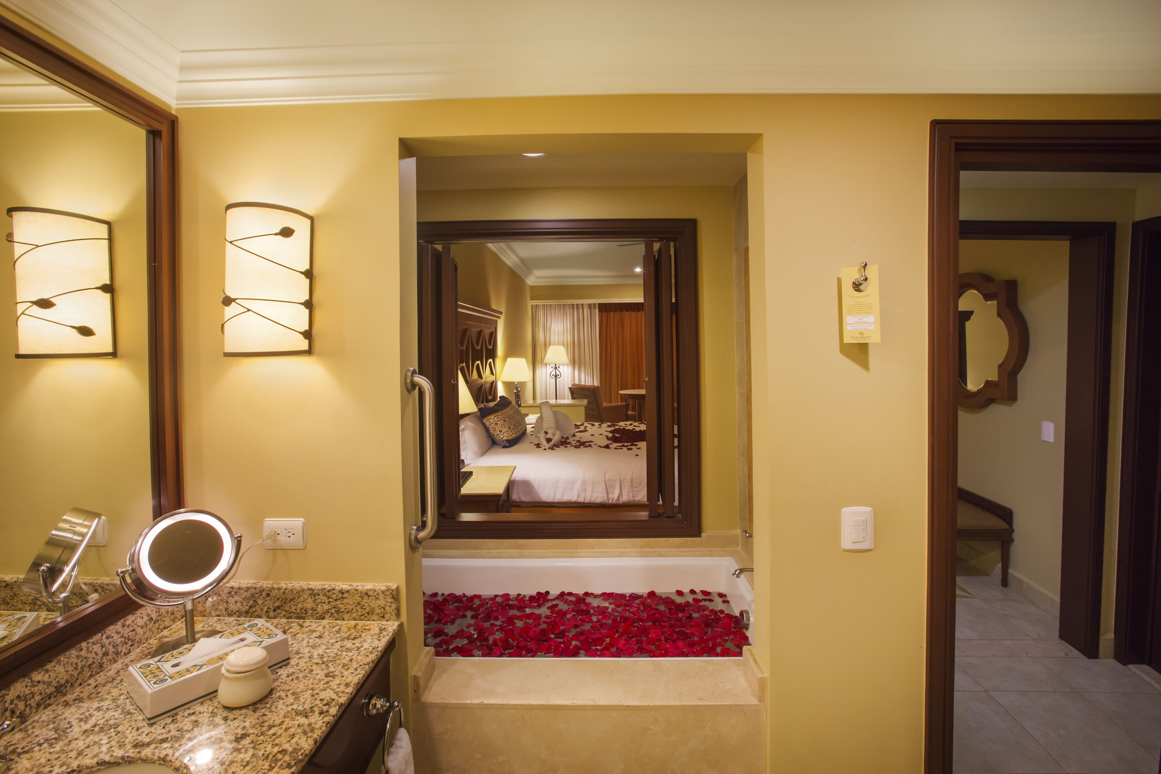 A hotel bathroom with a bathtub filled with rose petals, a vanity with a mirror and lights, and a view into the adjacent bedroom.