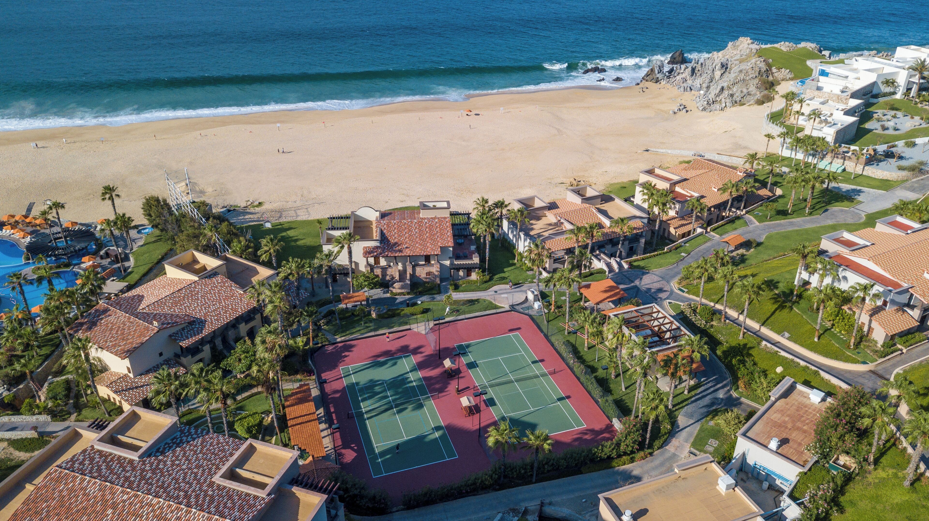 Vista aérea de un complejo turístico frente al mar, con playa, canchas de tenis y piscina.