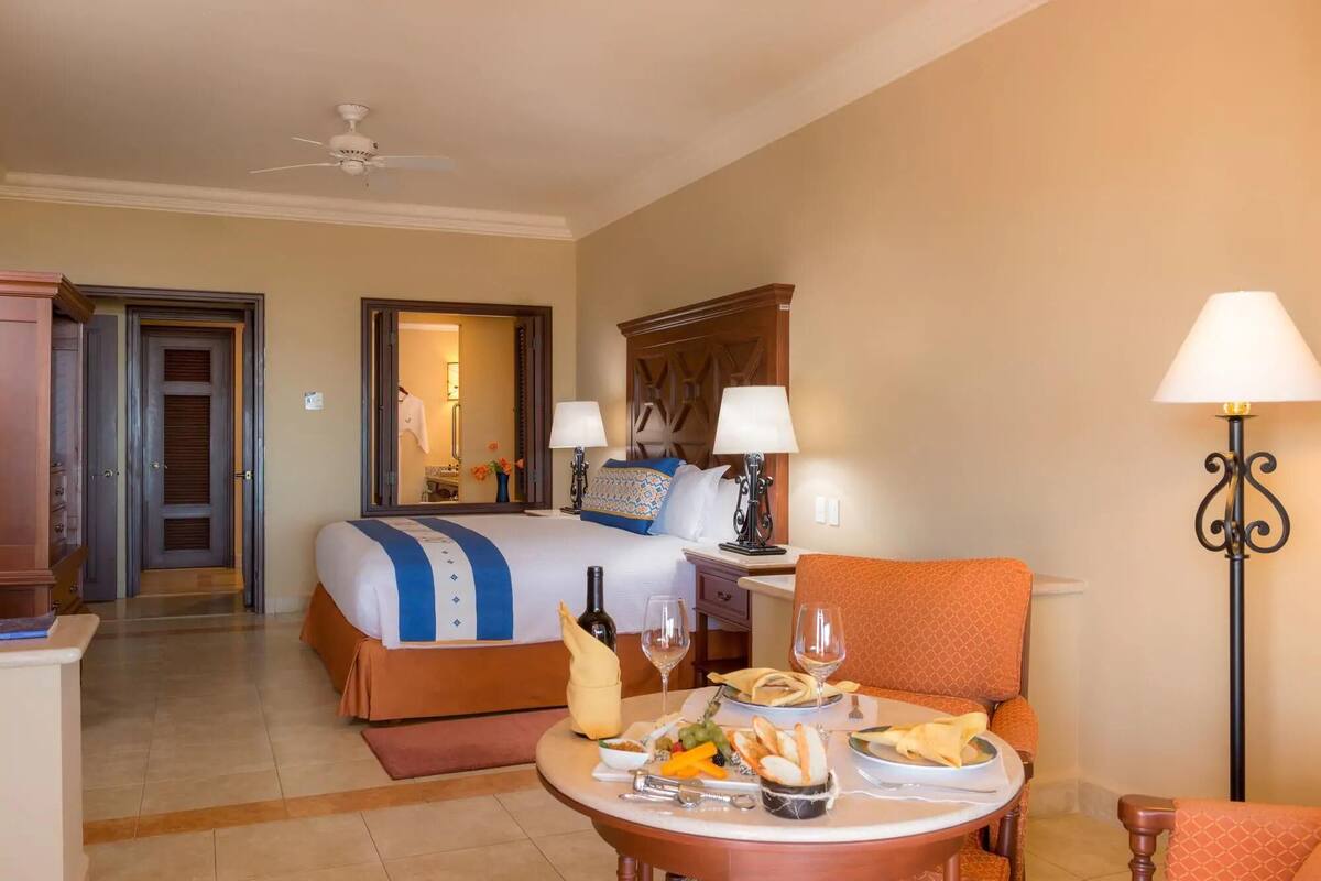 A hotel room with a bed, bedside tables, a dining table with food and drinks, a chair, and a lamp.