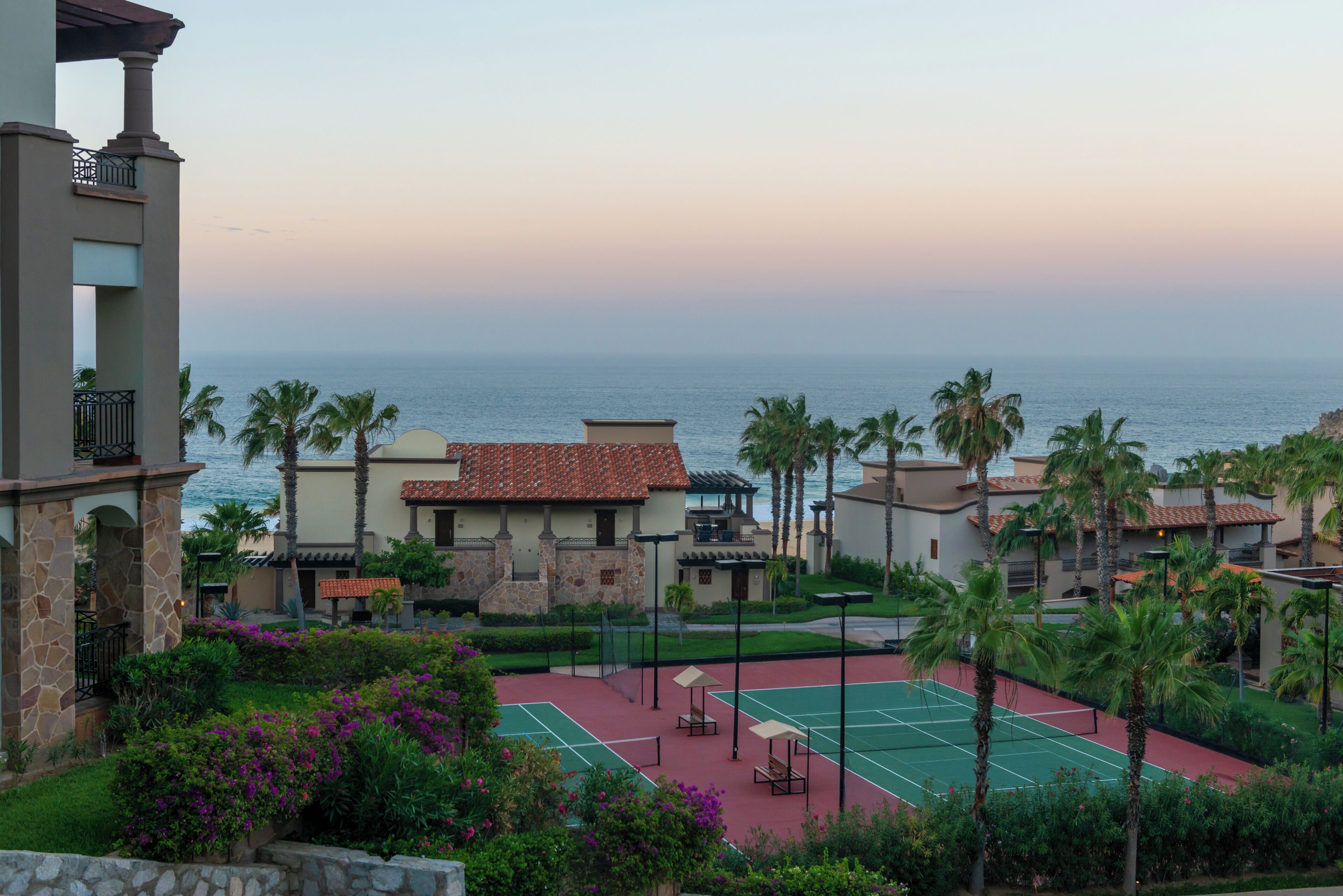 Un resort con cancha de tenis, palmeras y vista al océano.