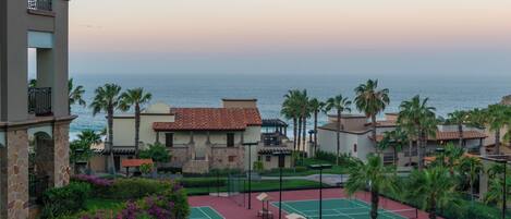 A resort with a tennis court, palm trees, and a view of the ocean.