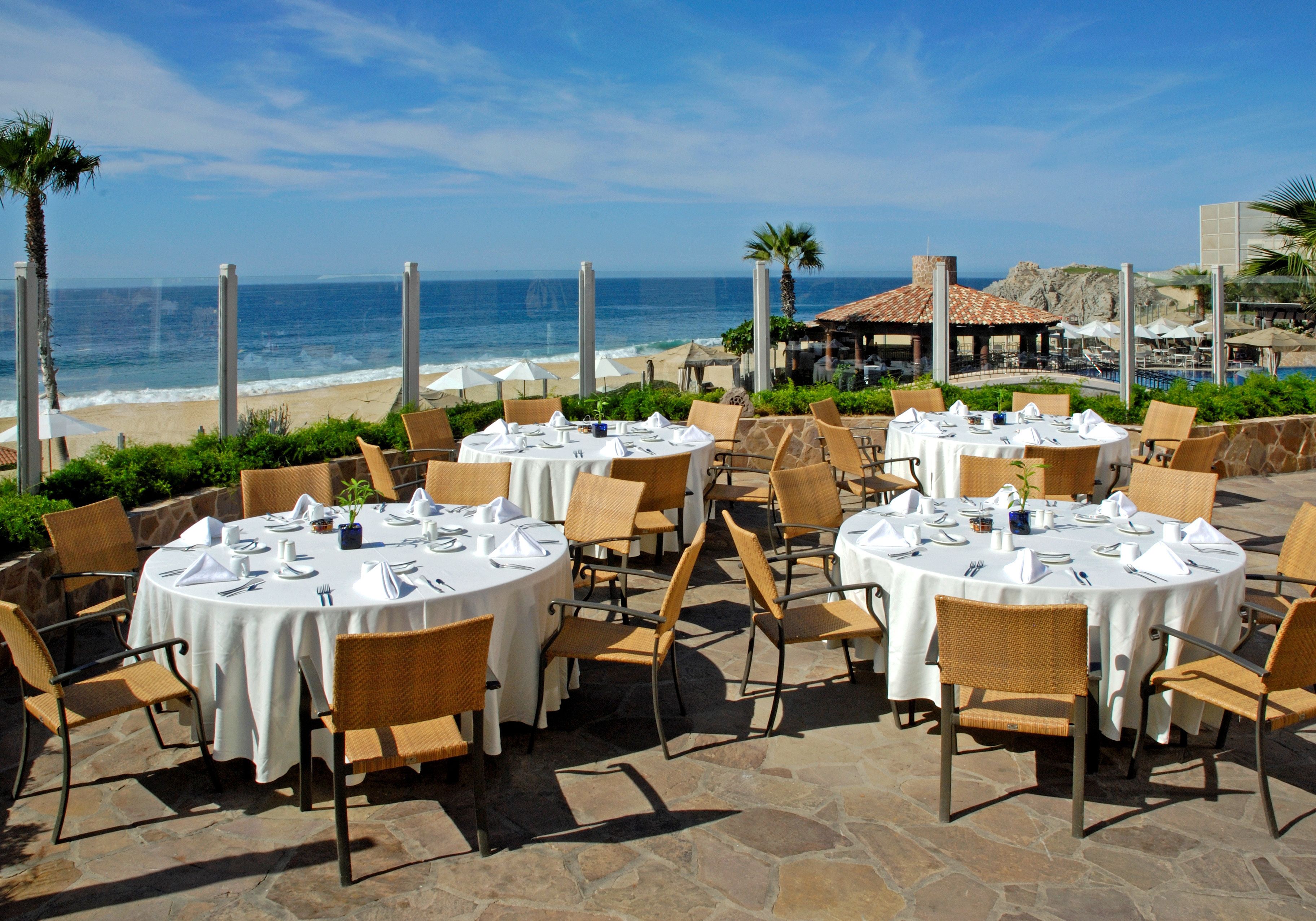 An outdoor dining area with round tables and wicker chairs, overlooking the ocean.
