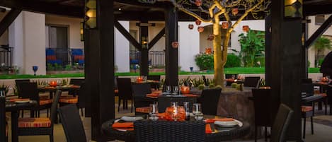 An outdoor dining area with wicker tables and chairs, a tree with hanging ornaments, and a view of a building in the background.
