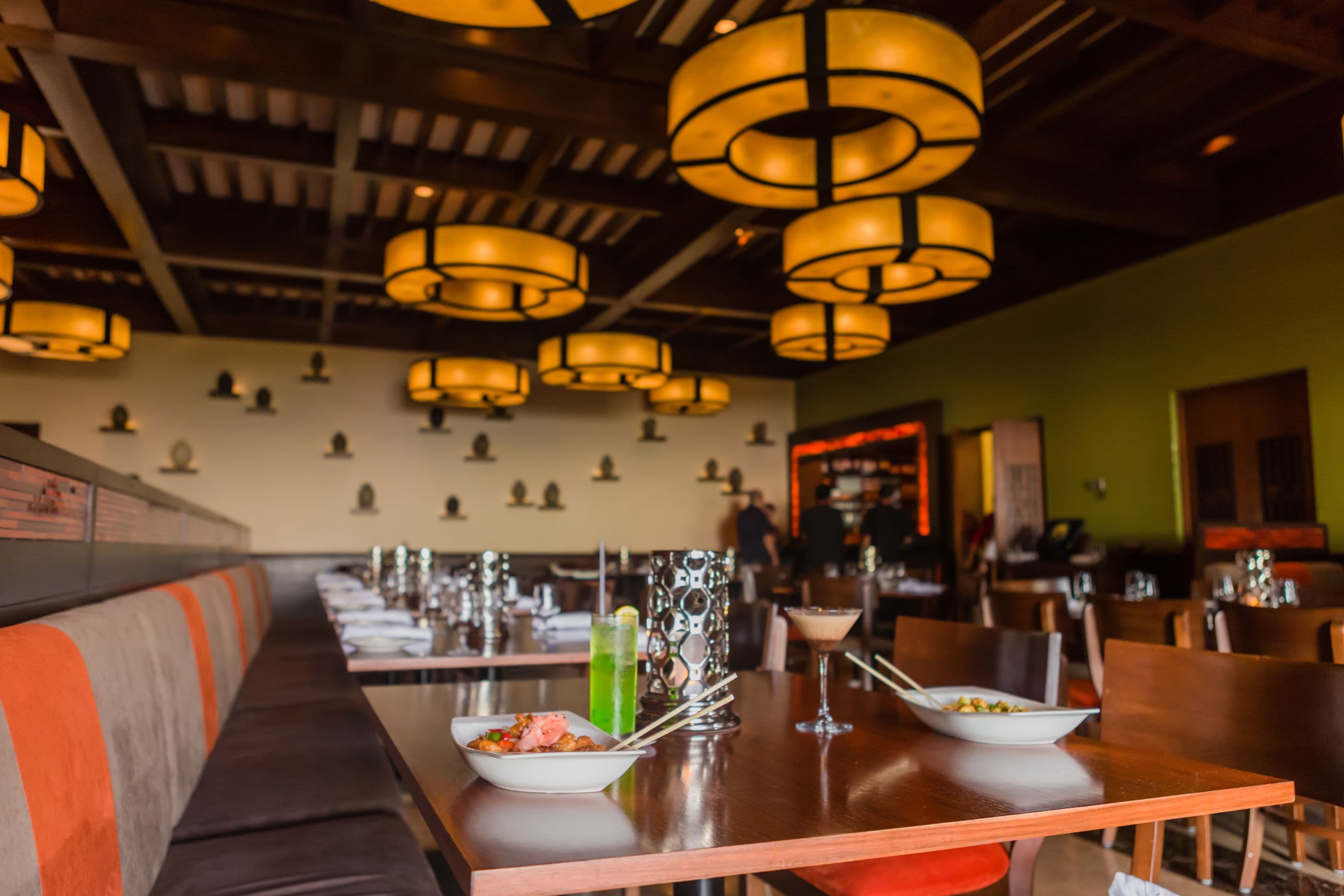 A restaurant interior with long communal tables, hanging pendant lights, and a green accent wall.