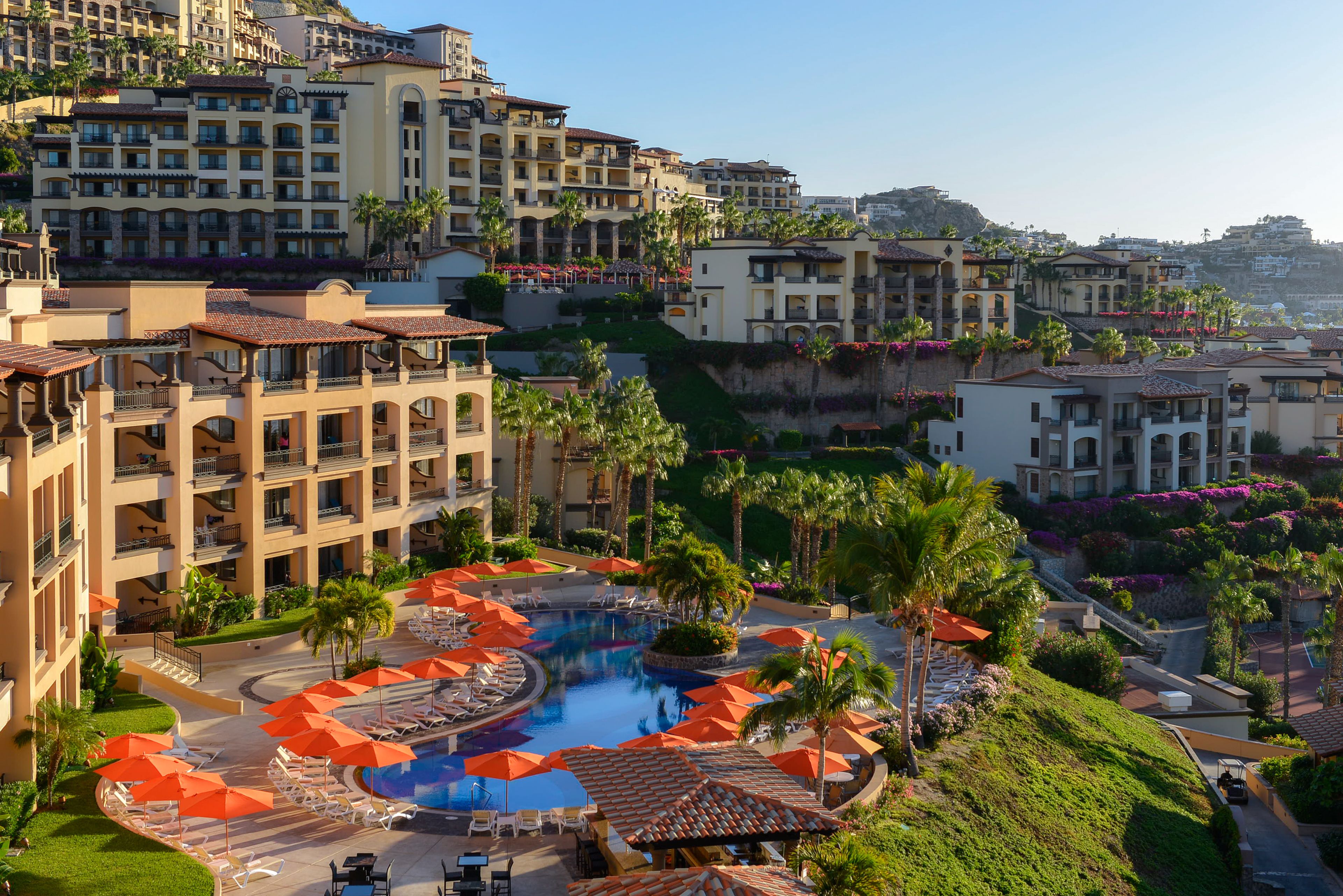 Un hotel con piscina rodeada de tumbonas y sombrillas, varios pisos con balcones y abundante vegetación.