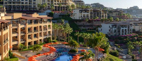 A hotel with a swimming pool surrounded by sun loungers and umbrellas, multiple floors with balconies, and lush landscaping.