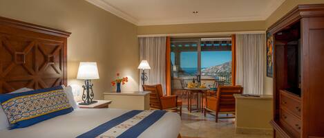 A hotel room with a large bed, a TV, a desk with a chair, a balcony with a view of the mountains, and a ceiling fan.