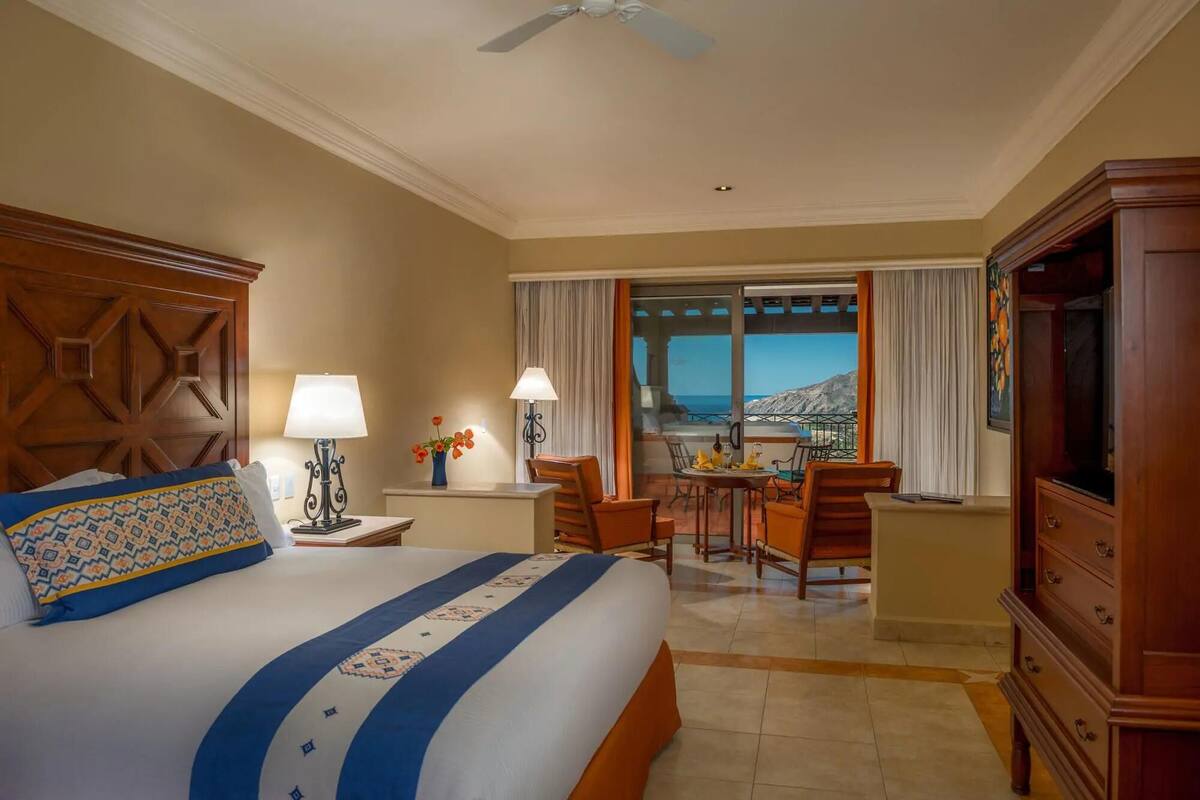 A hotel room with a bed, a desk with chairs, a television, and a view of the ocean.