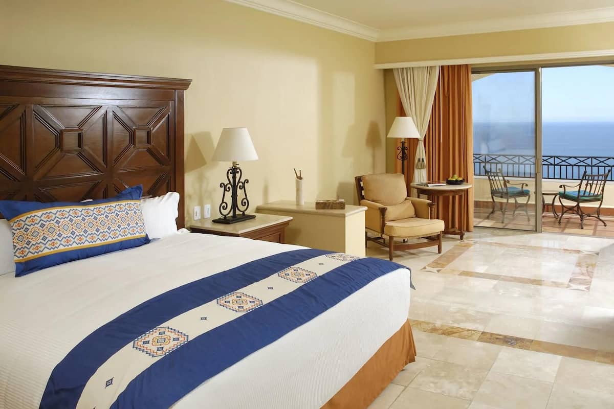 Habitación de hotel con una cama grande, una silla, una mesita y un balcón con vista al océano.