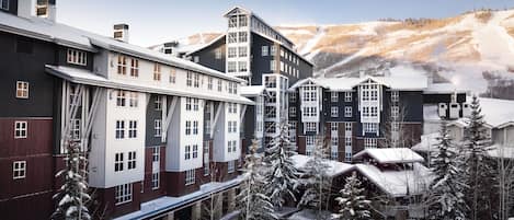 A multi-story hotel with a modern architectural design, featuring a mix of dark and light facades, and surrounded by snow-covered trees and a mountainous backdrop.