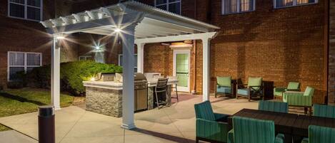 A covered patio area with outdoor furniture and a grill, adjacent to a brick building with windows.