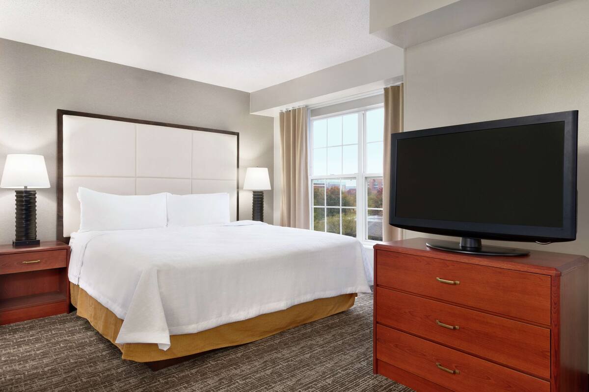 A hotel room with a large bed, two bedside tables with lamps, a television on a wooden dresser, and a window with curtains.