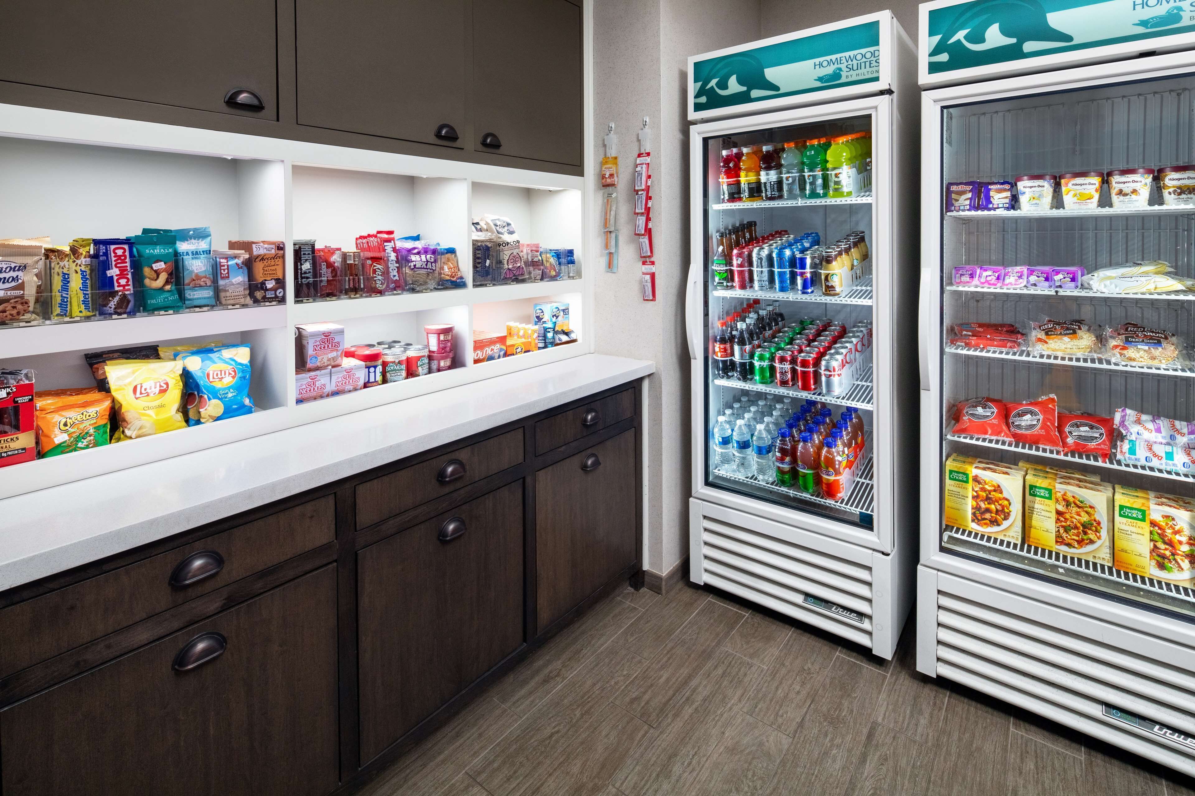 A kitchenette with a white countertop, wooden cabinets, and two mini-fridges stocked with snacks, drinks, and condiments.