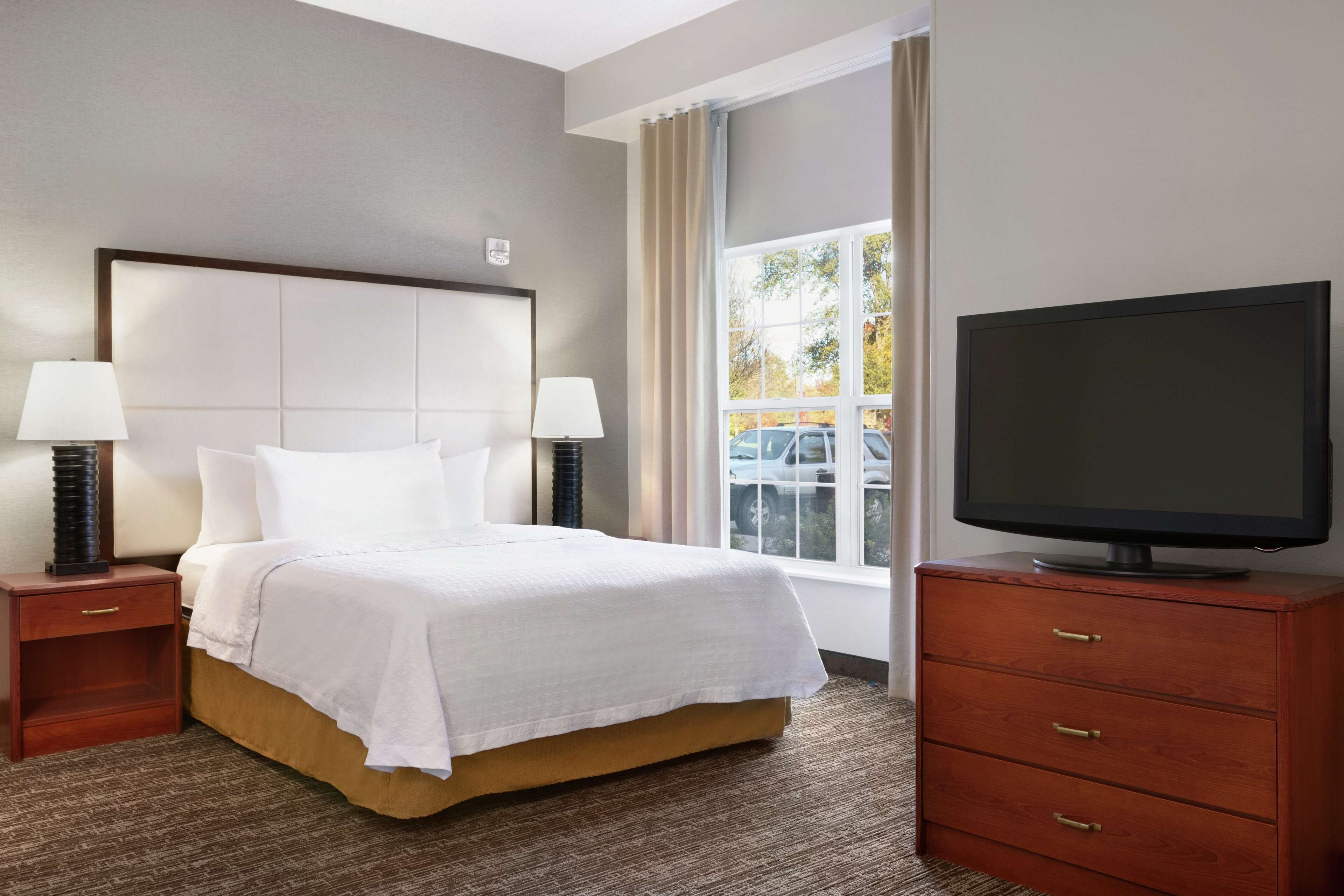 A hotel room with a bed, two bedside tables with lamps, a TV on a wooden cabinet, and a window with curtains.