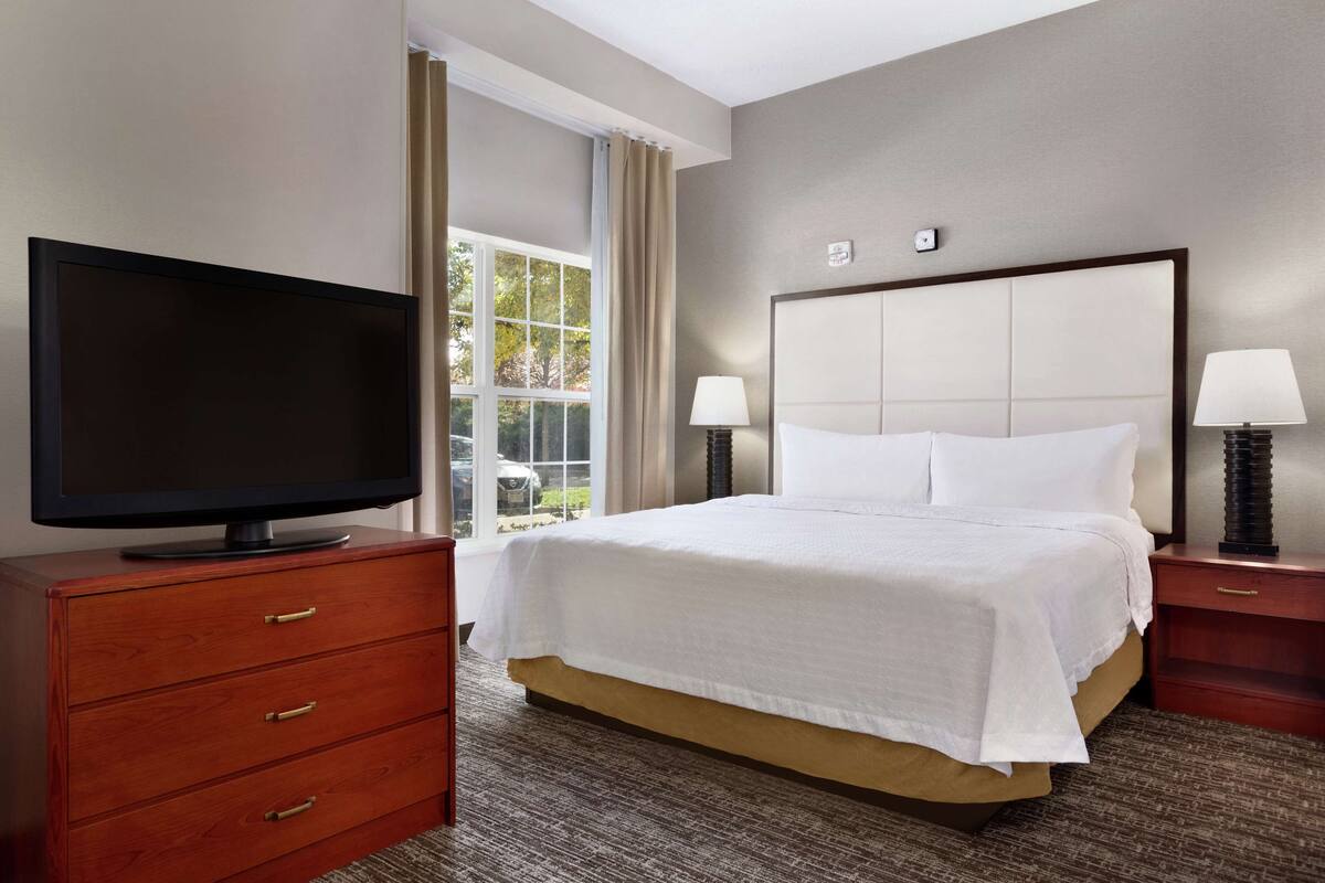 A hotel room with a bed, a television on a wooden dresser, two bedside lamps, and a window with curtains.