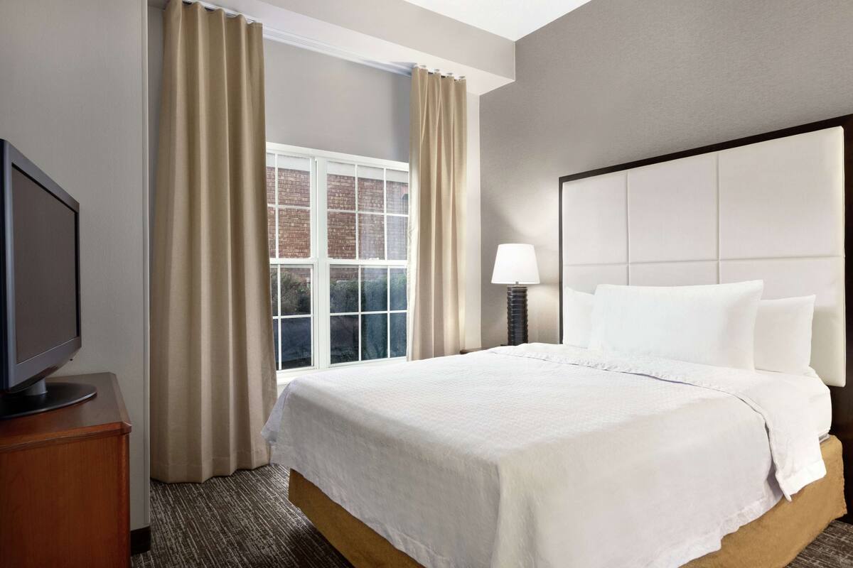 A hotel room with a large bed, a TV on a wooden stand, a window with curtains, and a nightstand with a lamp.