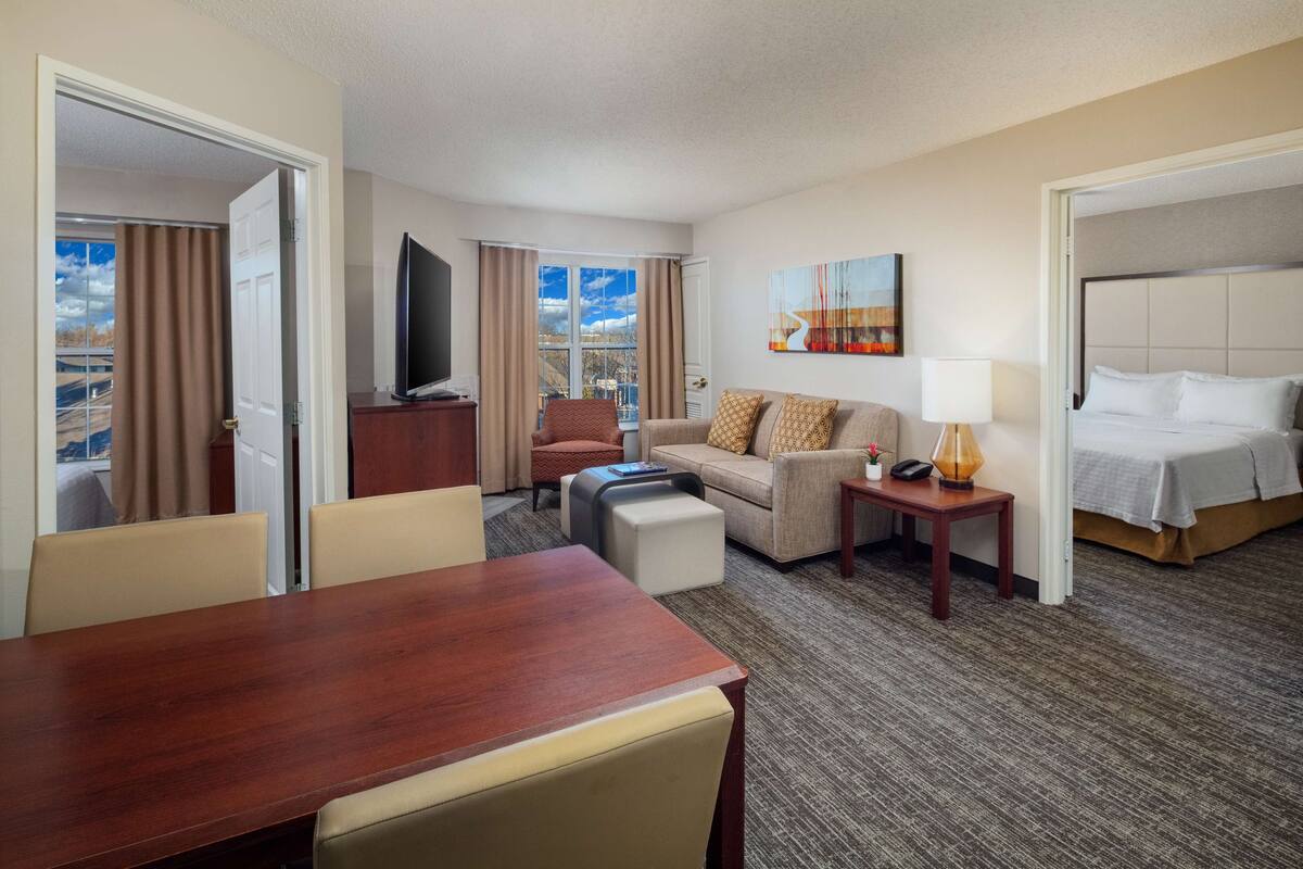 A hotel room with a wooden dining table, a sofa, a TV, a bed, and a view of the city.