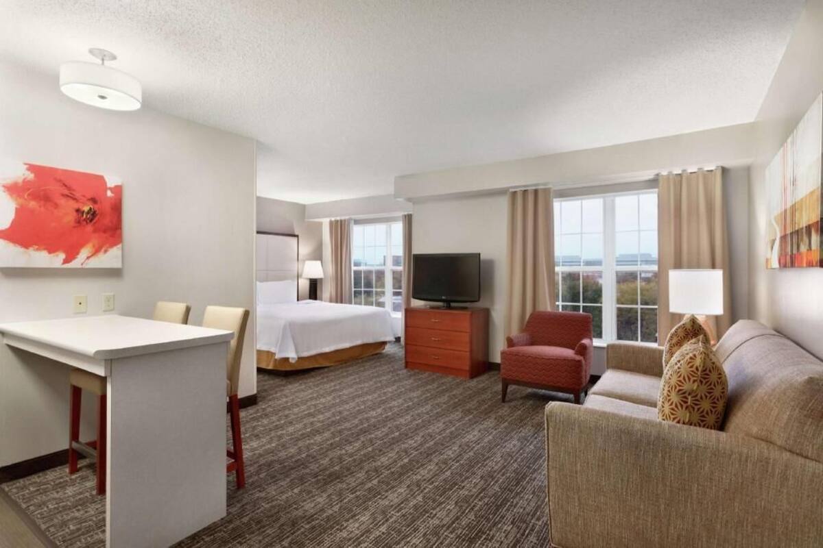 A hotel room with a bed, a TV, a desk, a chair, a sofa, and a window with curtains.