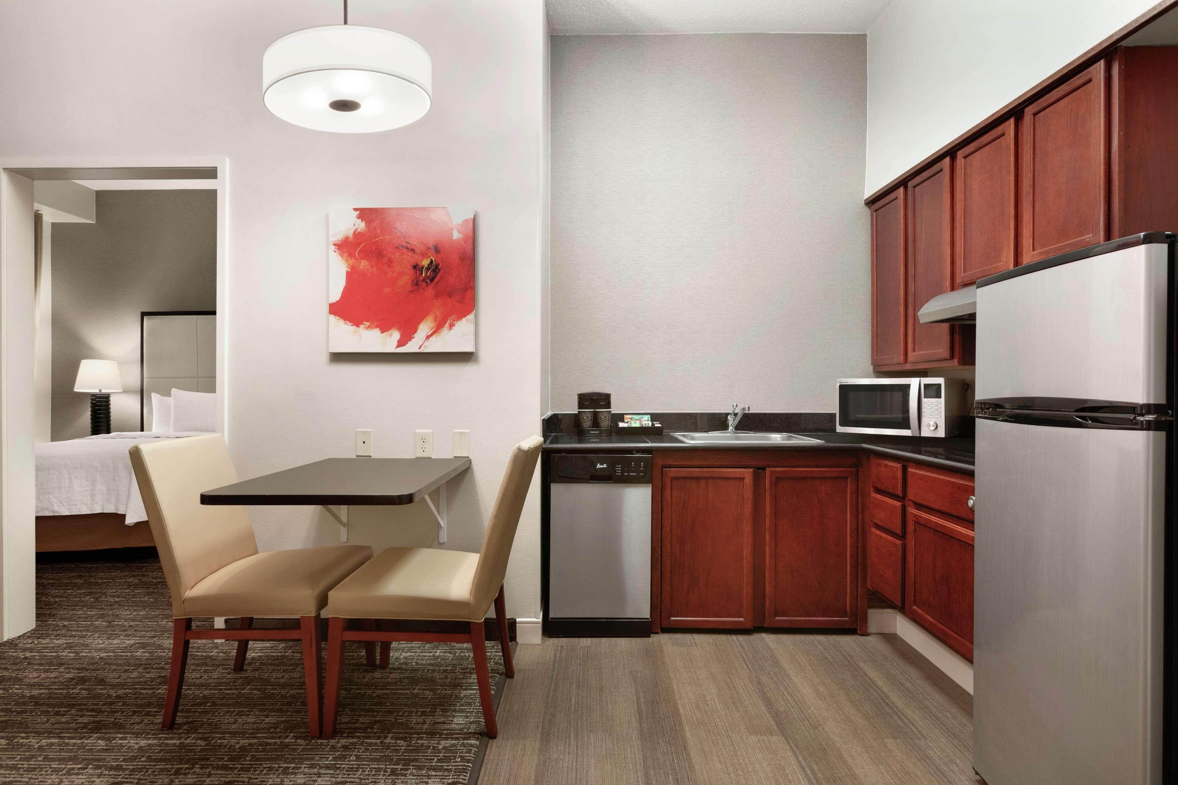 Una habitación de hotel compacta con kitchenette, una zona de comedor con mesa y sillas, y una cama con mesita de noche.