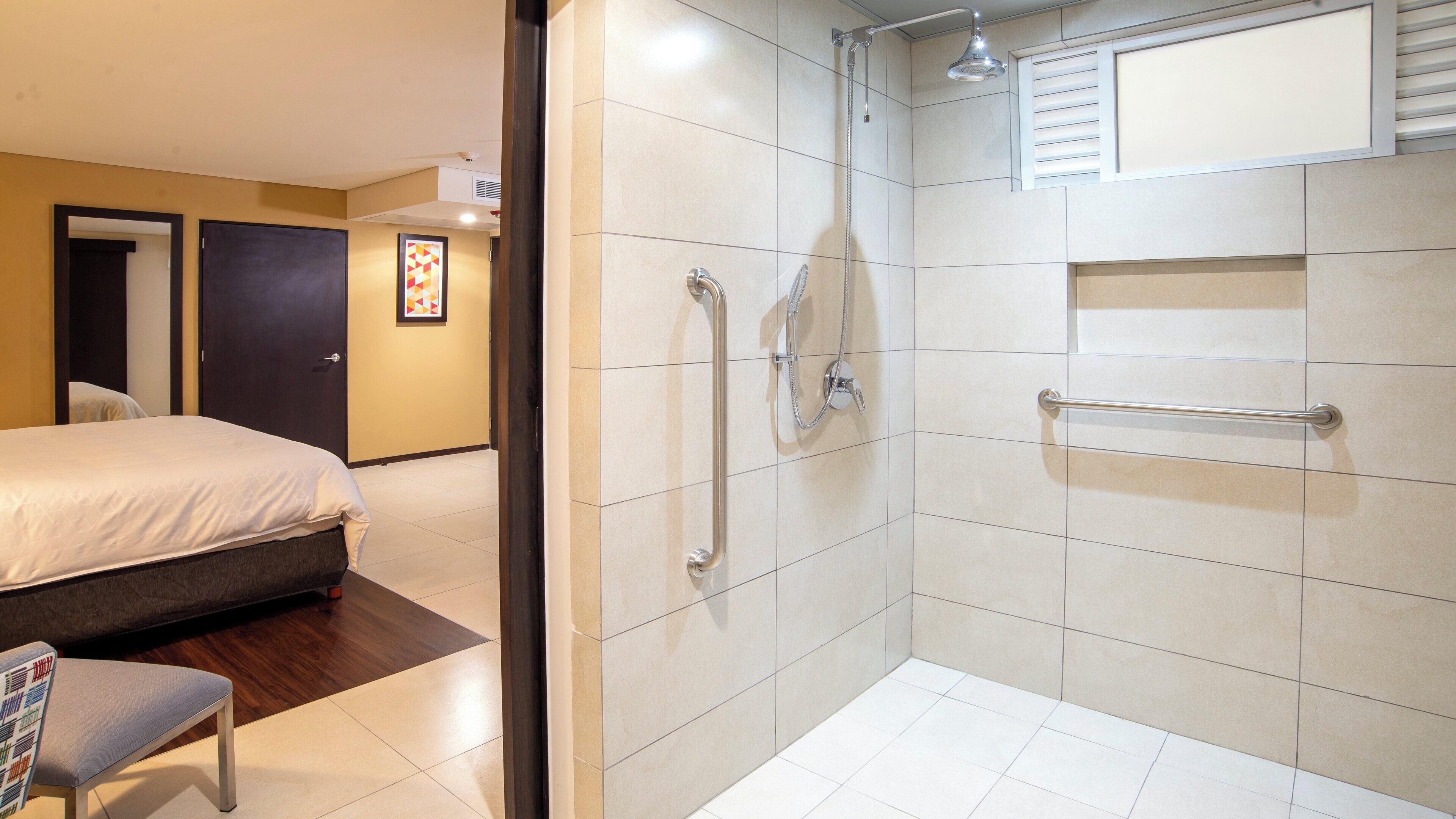 A hotel room with a bed, a bench, a shower with grab bars, and a towel rack.