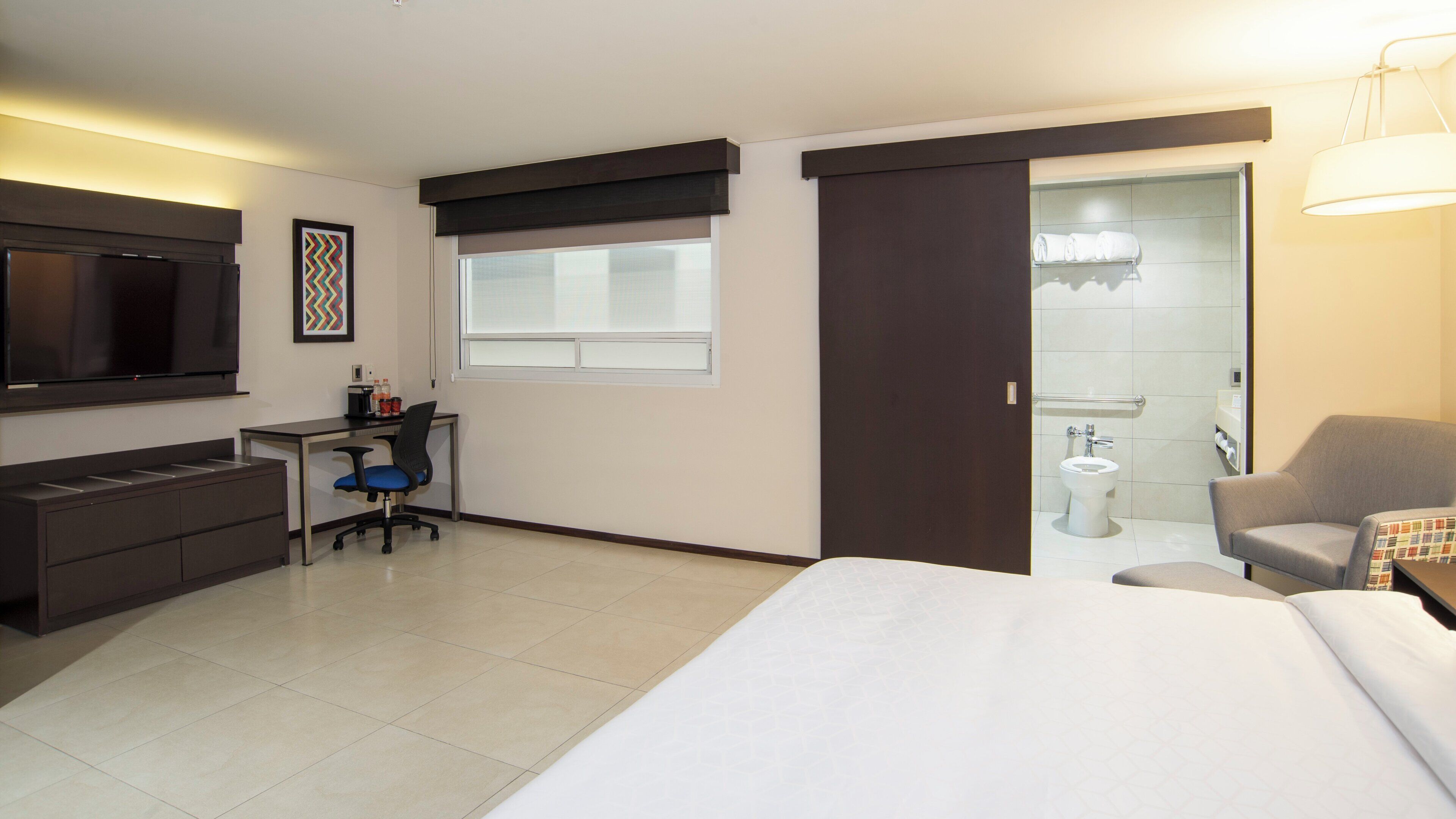 A hotel room with a bed, a desk with a chair, a television, a window, and a bathroom with a toilet and sink.