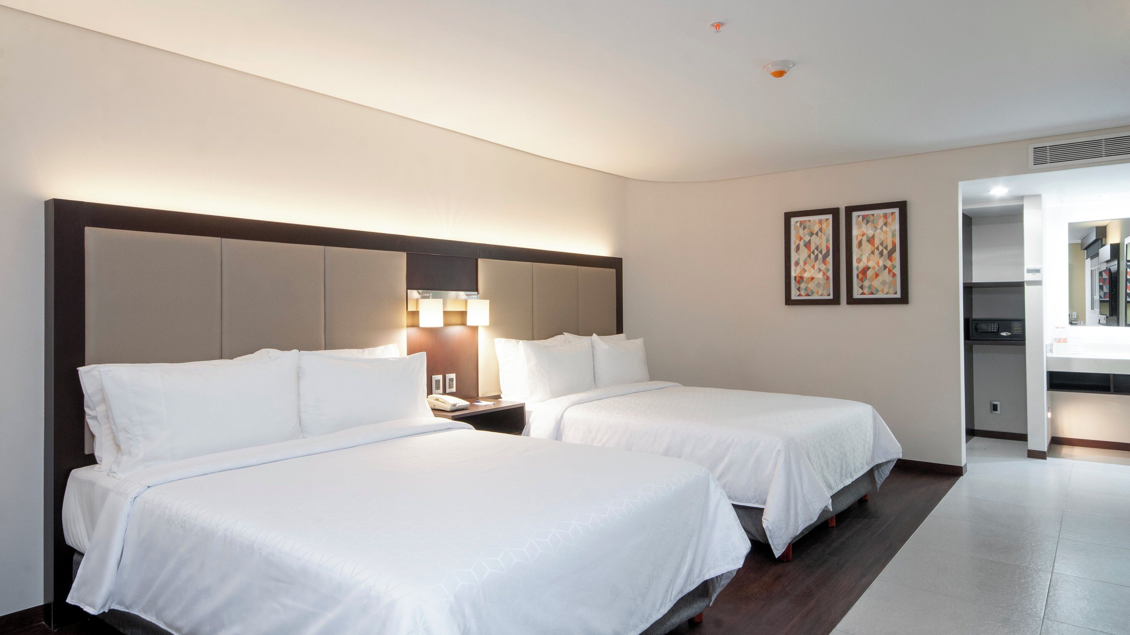 A hotel room with two beds, a nightstand, a lamp, and a kitchenette.