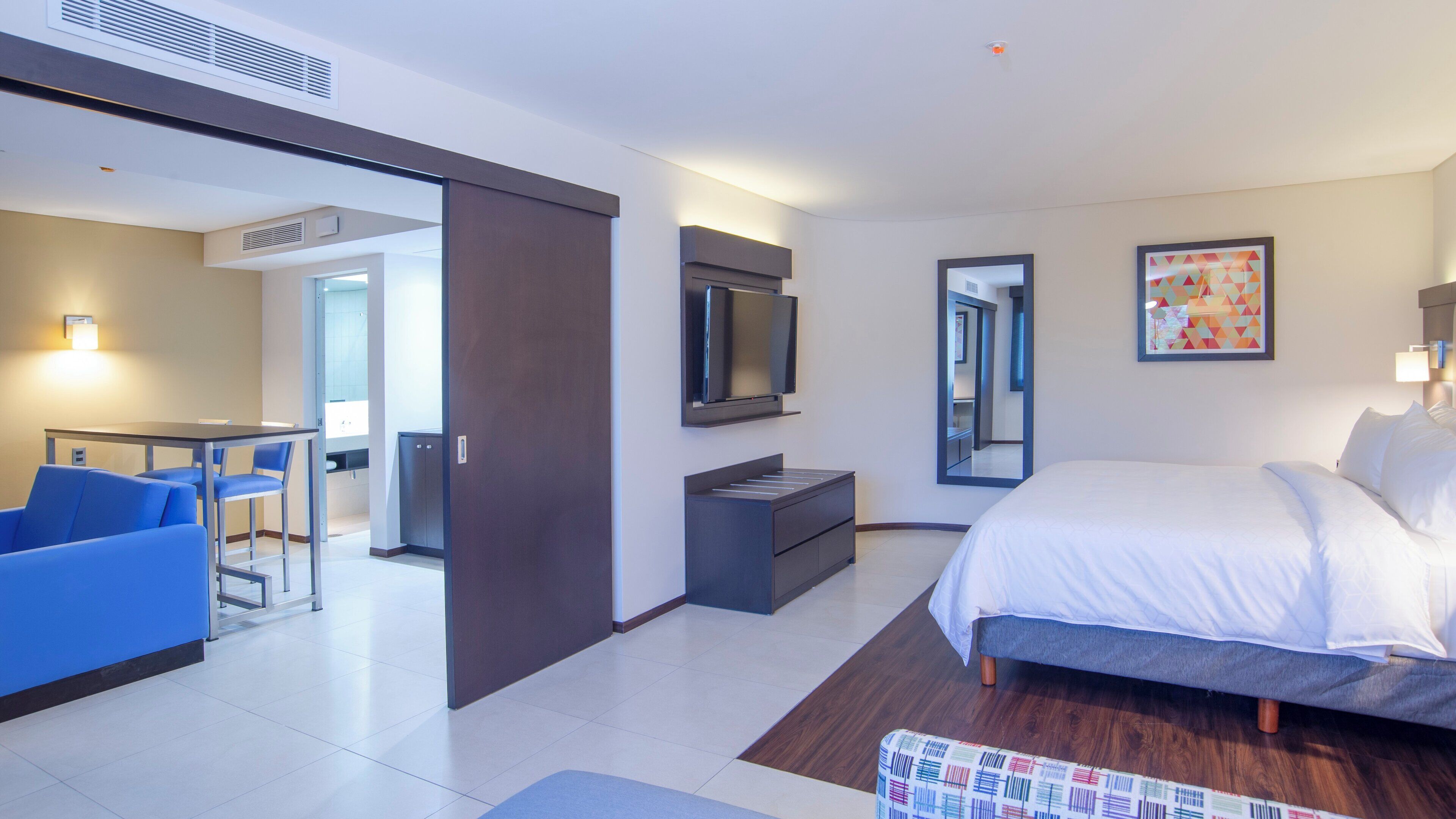 Ein modernes Hotelzimmer mit Bett, Schreibtisch, Stuhl, Fernseher und einem durch eine offene Tür einsehbarer Badezimmer.