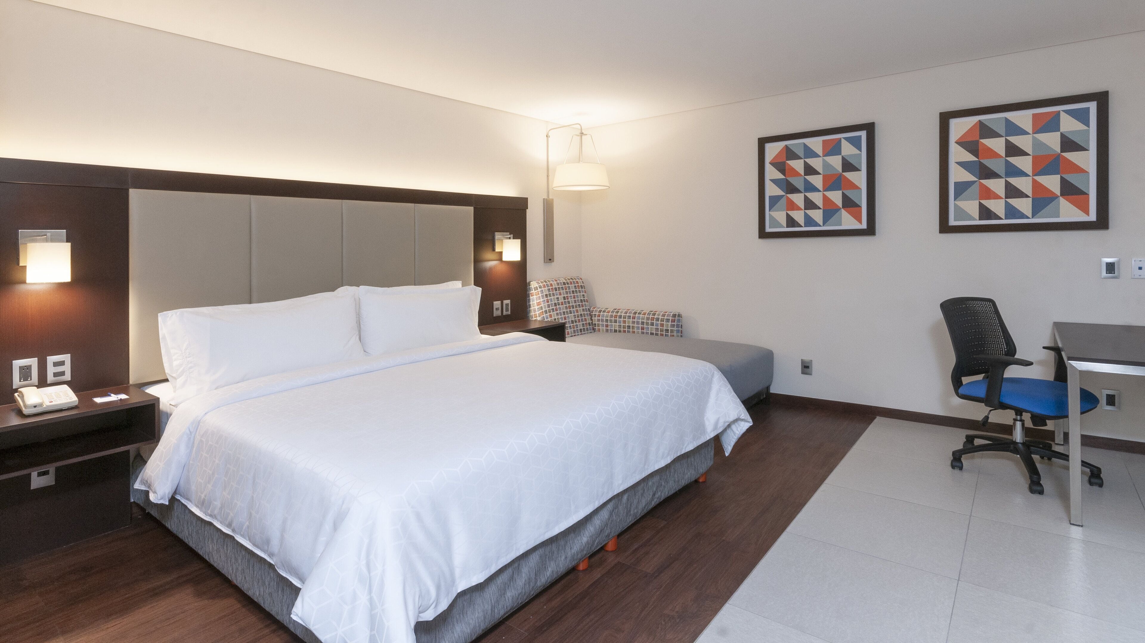 A hotel room with a large bed, a desk, and two framed geometric art pieces on the wall.