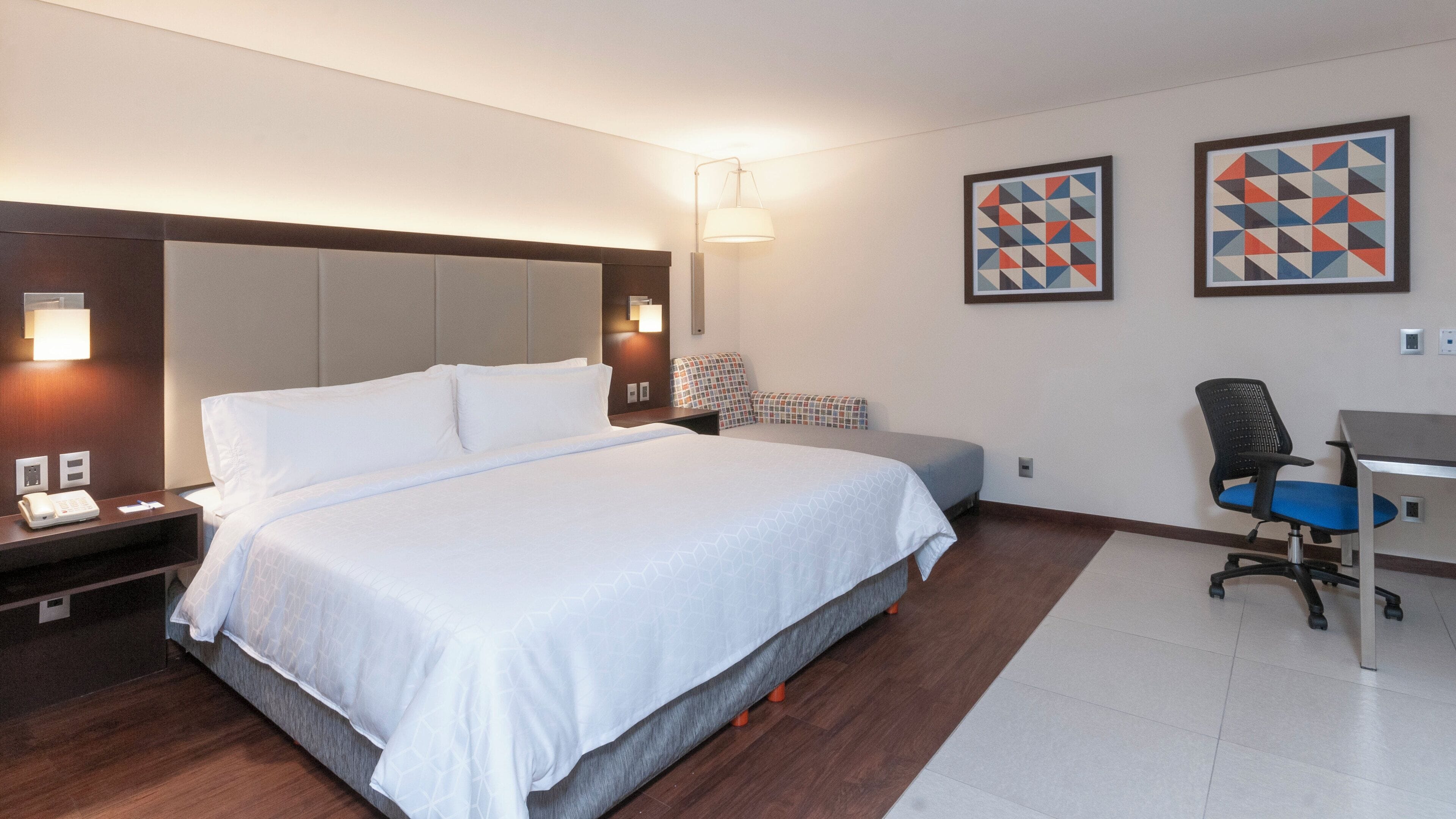 Une chambre d’hôtel avec un grand lit, des tables de chevet, un bureau et deux œuvres d’art géométriques encadrées accrochées au mur.