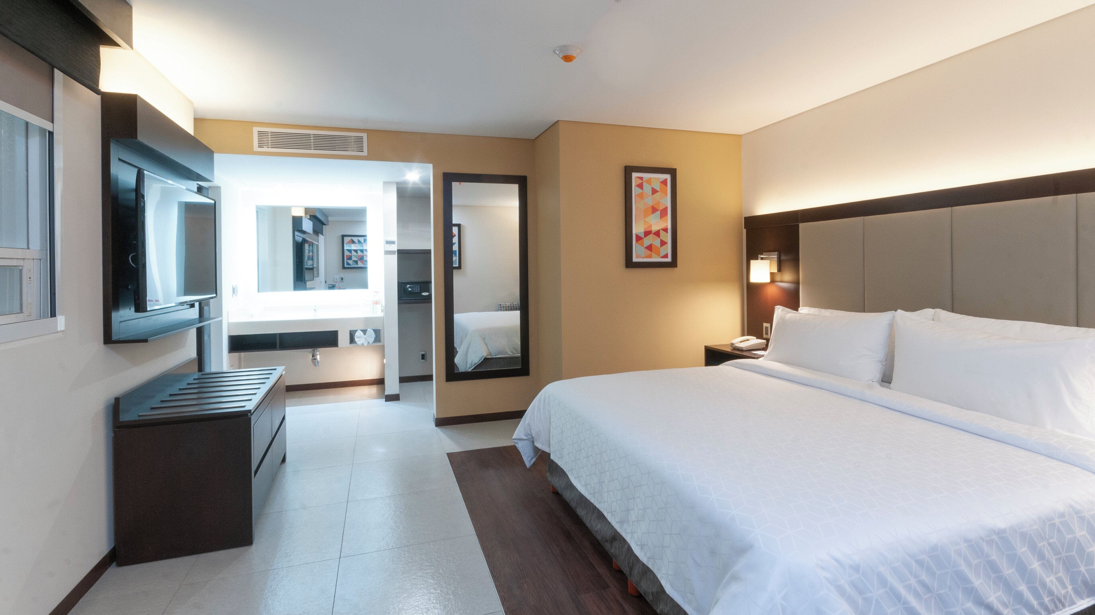 A hotel room with a large bed, a TV mounted on the wall, a bedside table, and a bathroom visible through an open door.