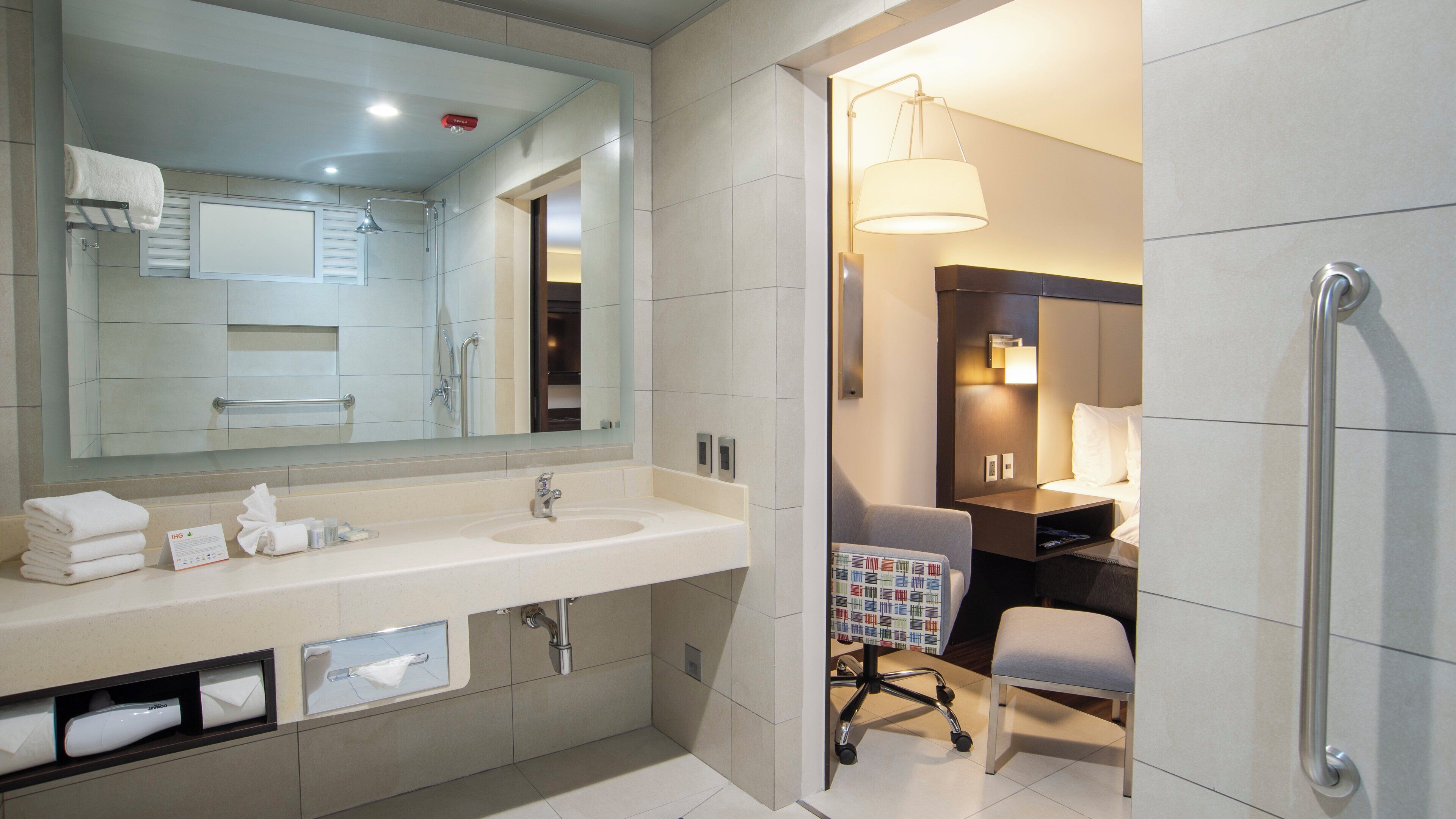 A modern bathroom with a large mirror, dual sinks, and a wheelchair-accessible chair.