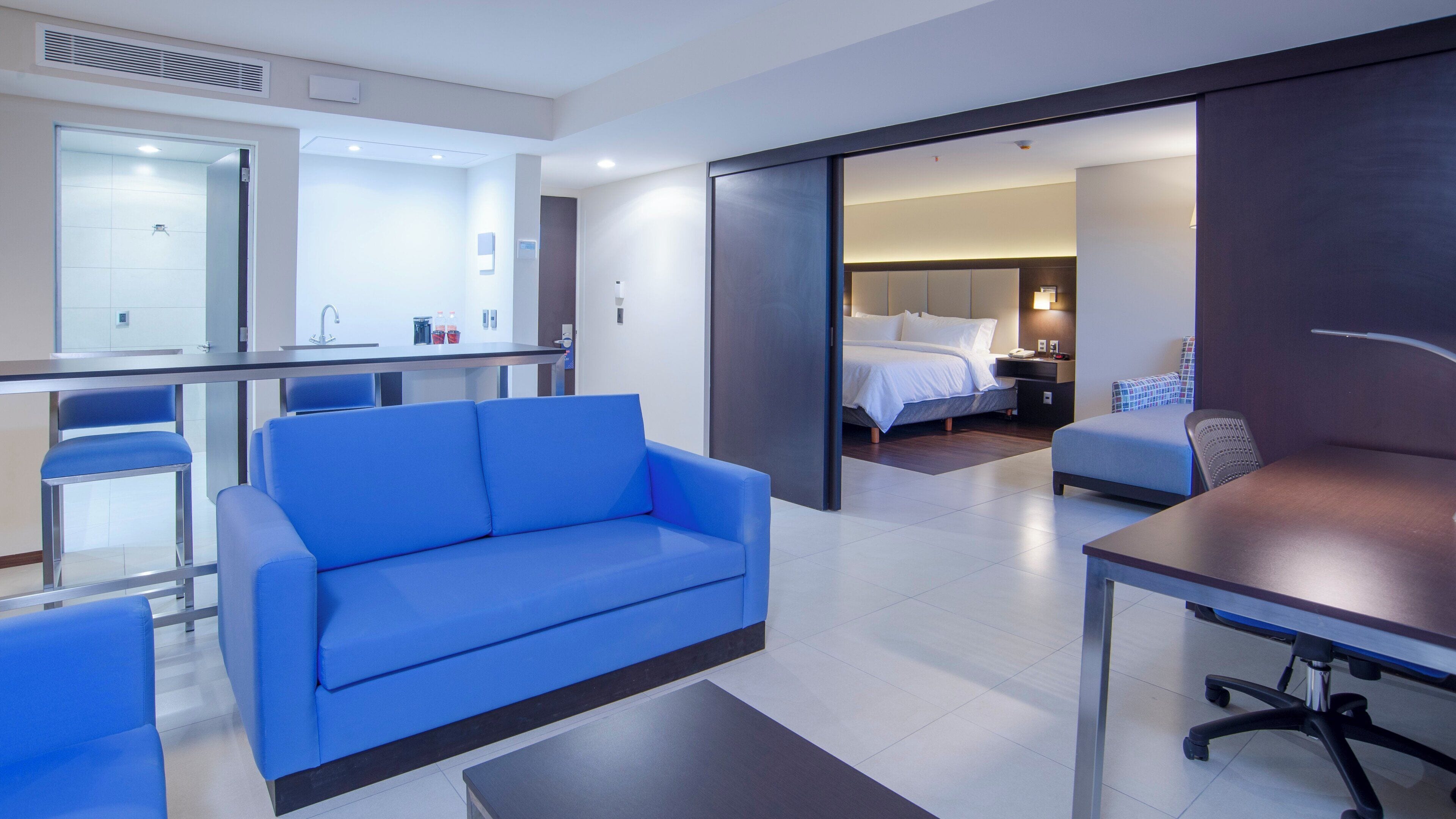 A modern hotel room with a blue sofa, a dining table, a separate bedroom with a bed, and a bathroom visible through an open door.