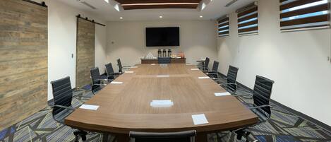 A conference room with a large wooden table, black chairs, a flat-screen TV, and a patterned carpet.