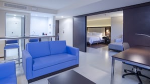 Habitación de hotel moderna con sofá azul, mesa de comedor, zona de cocina y dormitorio independiente con cama y mesita de noche.