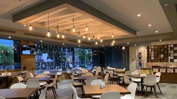 Un restaurante moderno con varias mesas y sillas, ventanas amplias y un techo con vigas a la vista y luces colgantes.