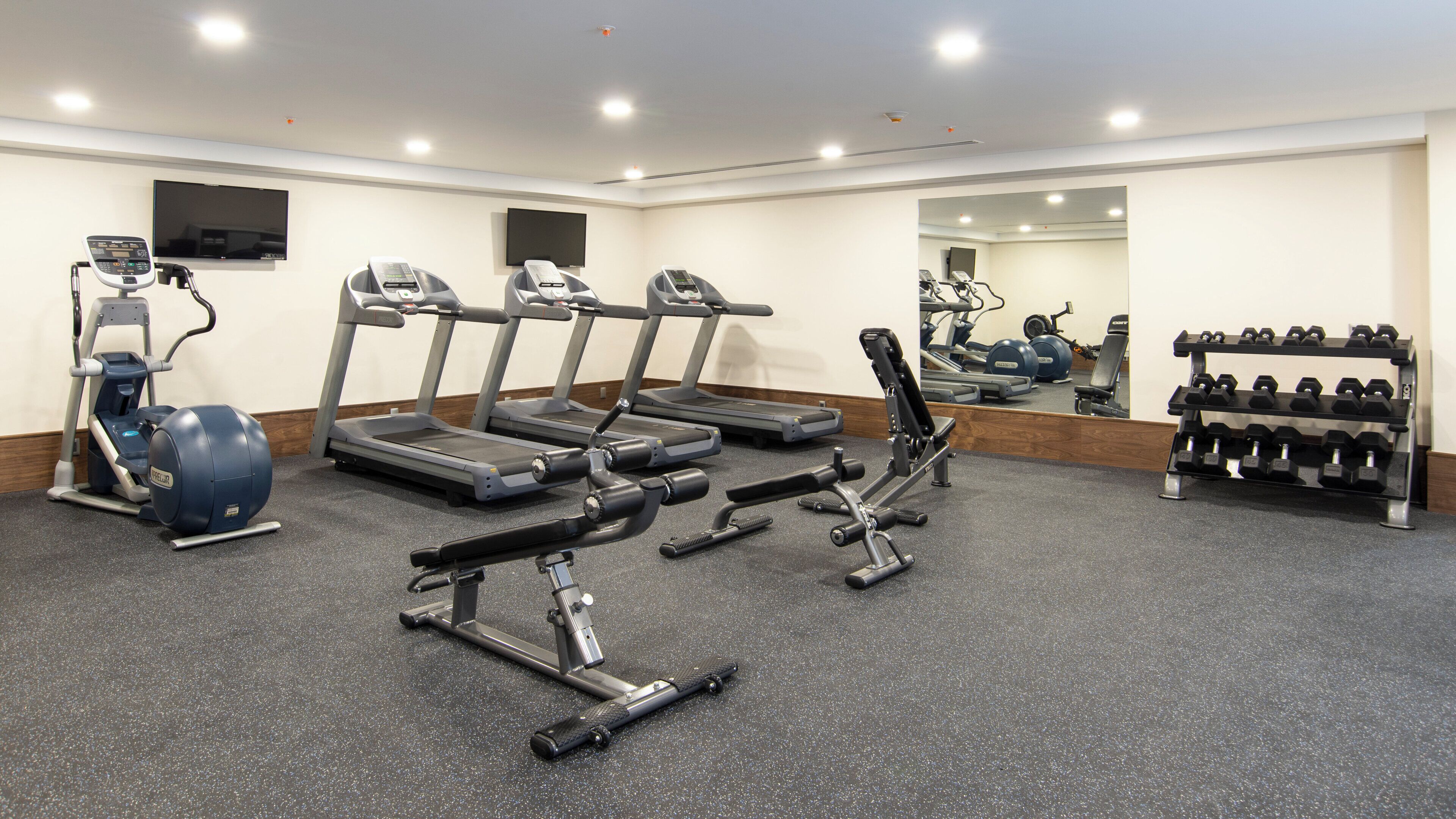 Uma academia com esteiras, elípticos, um banco de musculação e uma barra de halteres.
