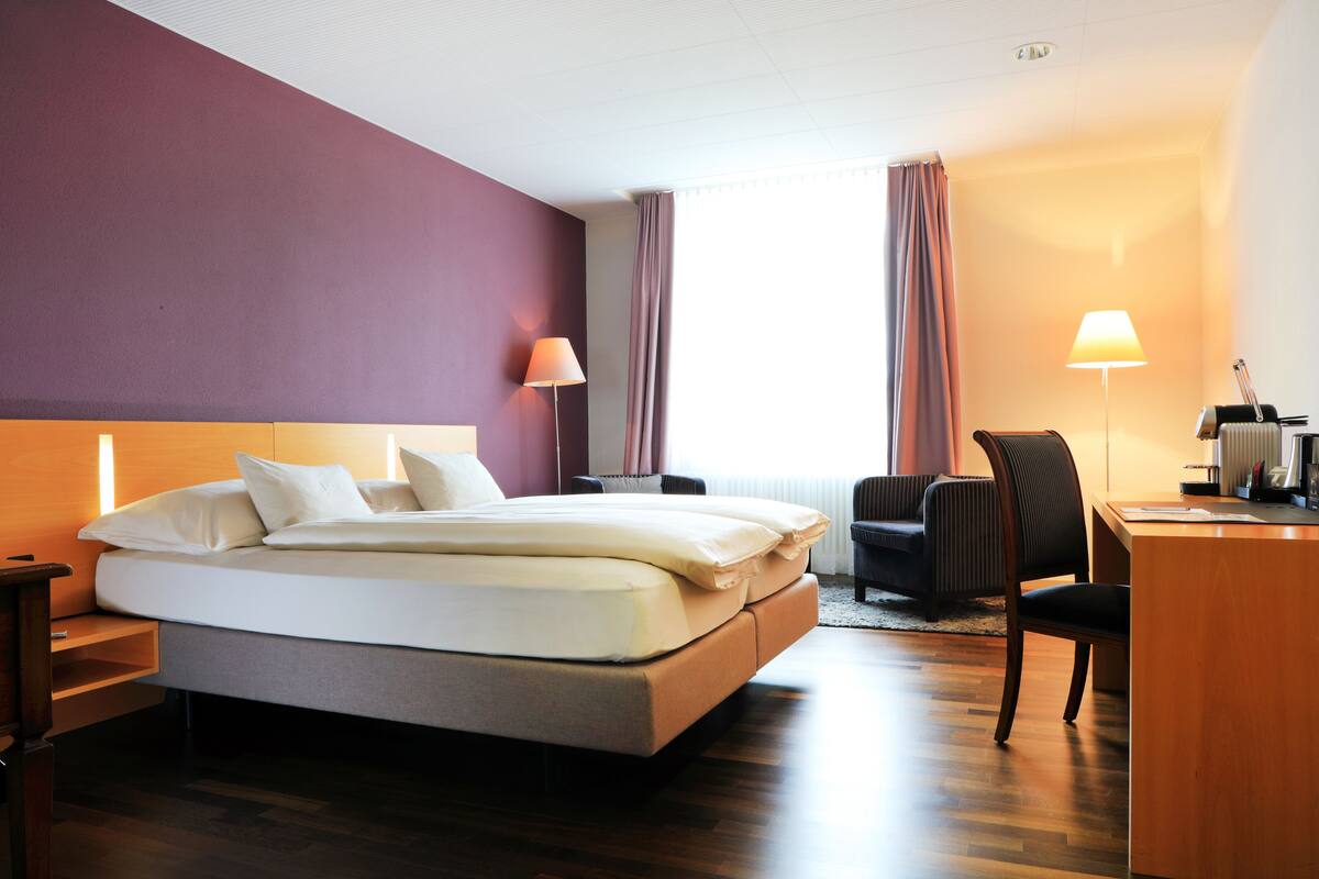 Ein Hotelzimmer mit Bett, Schreibtisch, Stuhl, Lampe und Blick ins Freie.