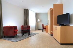A modern hotel room with a flat-screen TV, a red armchair, a sofa, a small table, and a lamp.